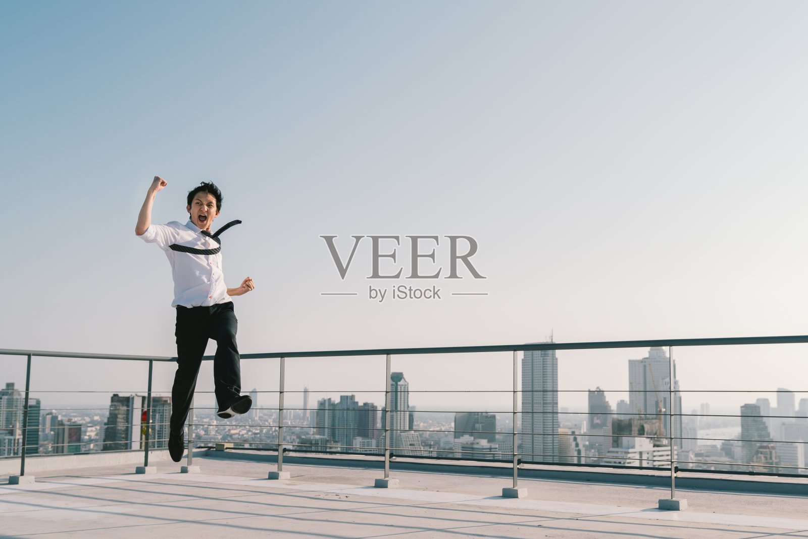 年轻英俊的亚洲商人跳高，庆祝成功获胜的姿态在建筑屋顶。工作、工作或成功的商业理念。城市景观背景与拷贝空间在阳光明媚的蓝天照片摄影图片