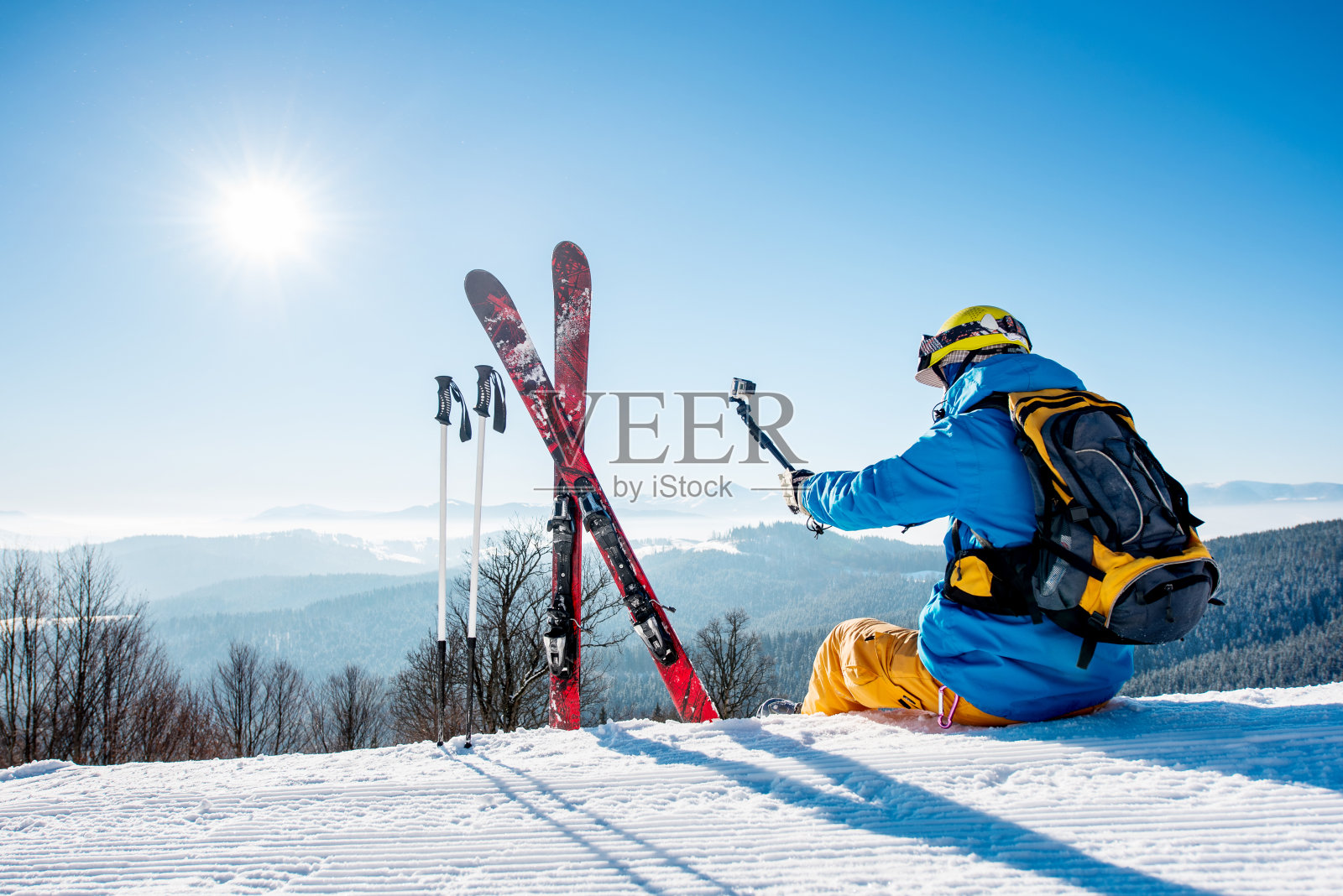一个滑雪者与背包和滑雪设备坐在山顶的雪自拍与他的相机在自拍杆上休息后在冬季度假村滑雪照片摄影图片