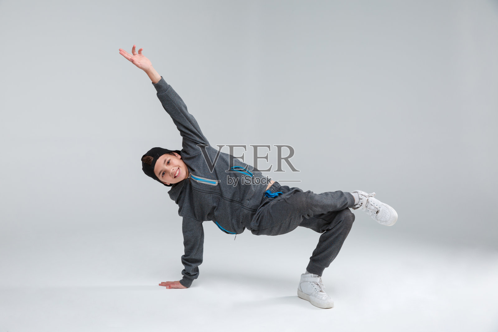 一个快乐的小男孩穿着灰色的运动服，积极地用一只胳膊和一条腿站着跳舞。灰色的背景。照片摄影图片