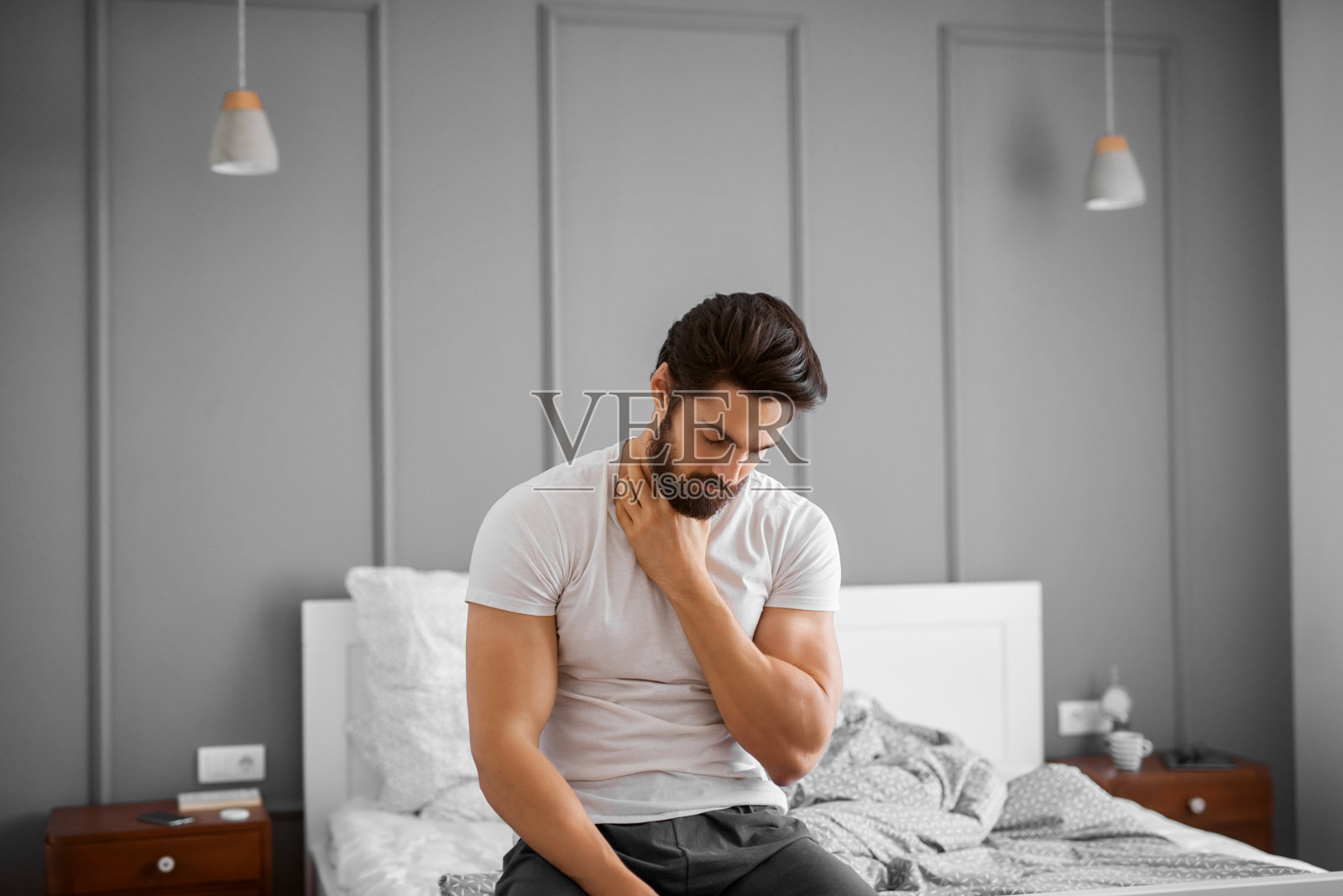 一幅愤怒的、肌肉发达、蓄胡须的英俊男子的肖像，他喉咙痛，坐在家里的床沿上。照片摄影图片