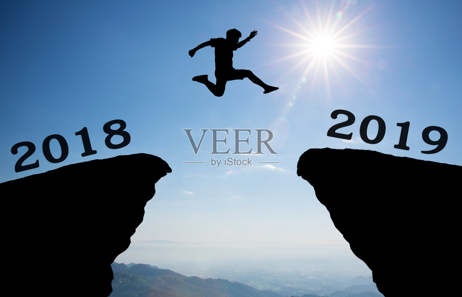 一个年轻的男子跳跃在2018年和2019年之间越过太阳，穿过山沟上剪影黄昏多彩的天空。照片摄影图片