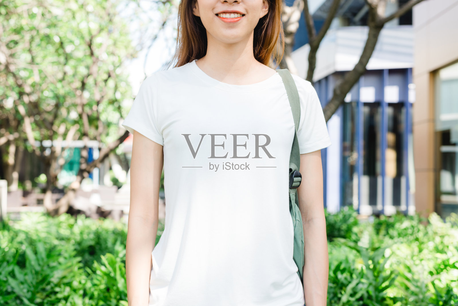 亚洲嬉皮女孩棕色长发在白色空白t恤站在街道中央。一位身着街头服饰的女性站在绿色的城市背景上。为文本或设计留出模拟空间。照片摄影图片