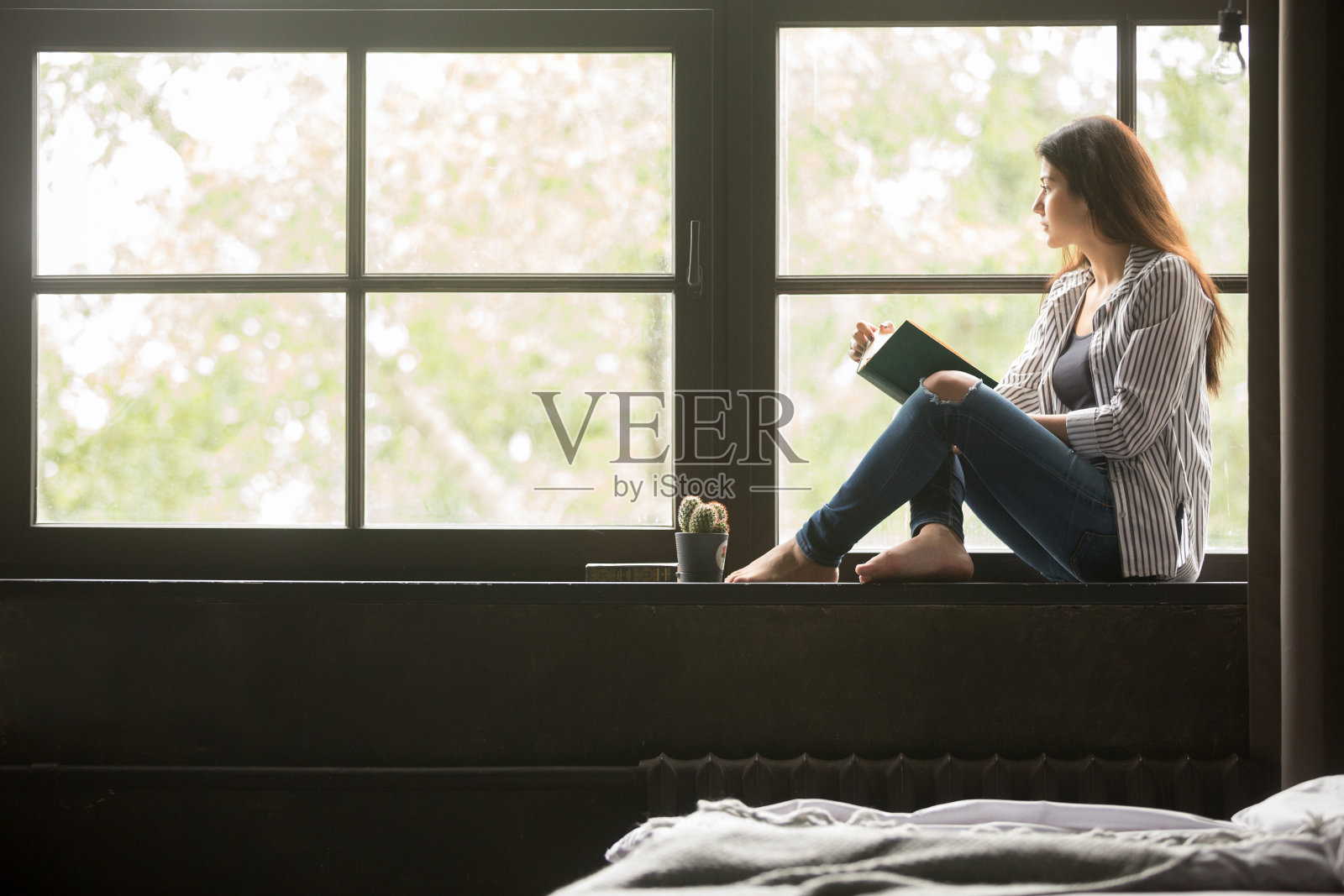 Pensive dreamy girl holding book sitting on sill at home照片摄影图片