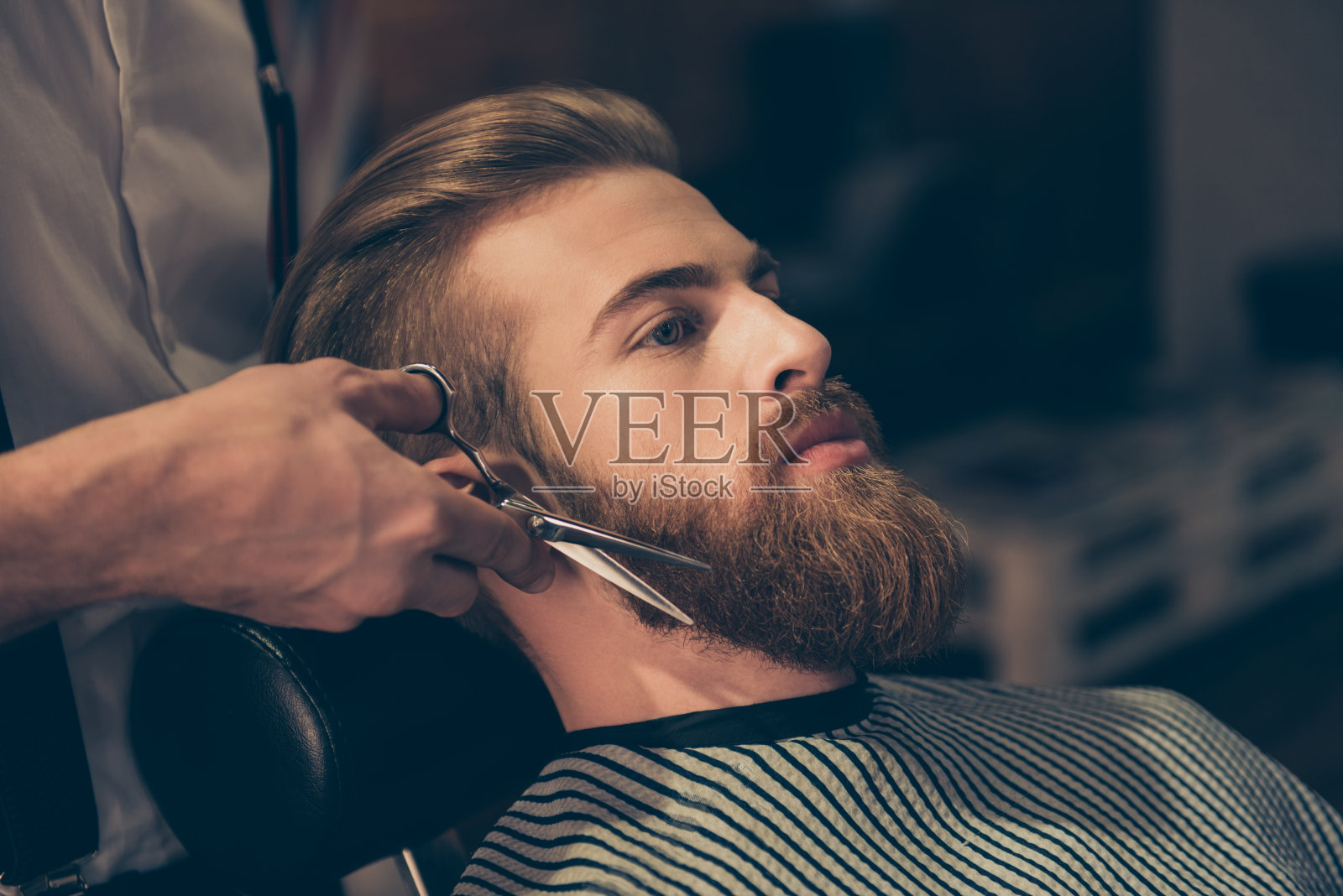 近距离的理发师的工作，一个有吸引力的年轻人在理发店。他正在用剪刀修剪红胡子照片摄影图片