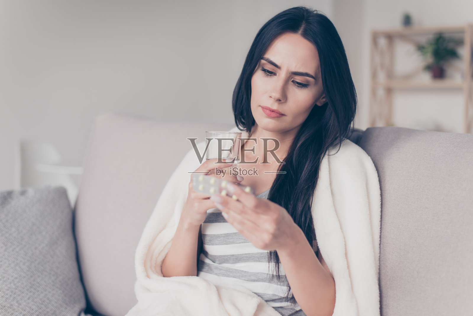 画像中，一个患病的年轻女子坐在家里，手里拿着一杯水，读着关于药物的描述照片摄影图片