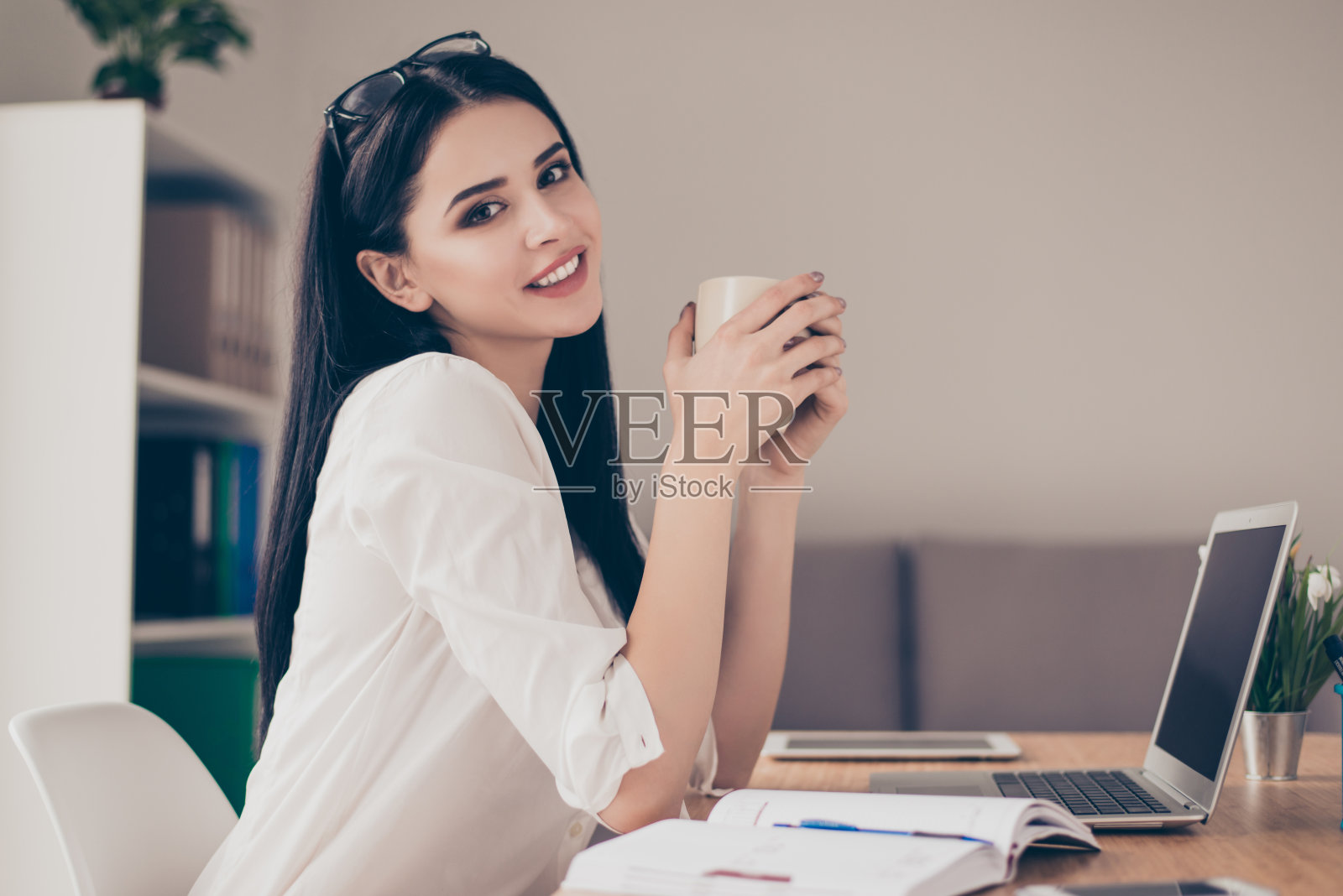 快乐美丽可爱的年轻轻松的女商人坐在桌子上休息，拿着一杯茶在灯光办公室照片摄影图片