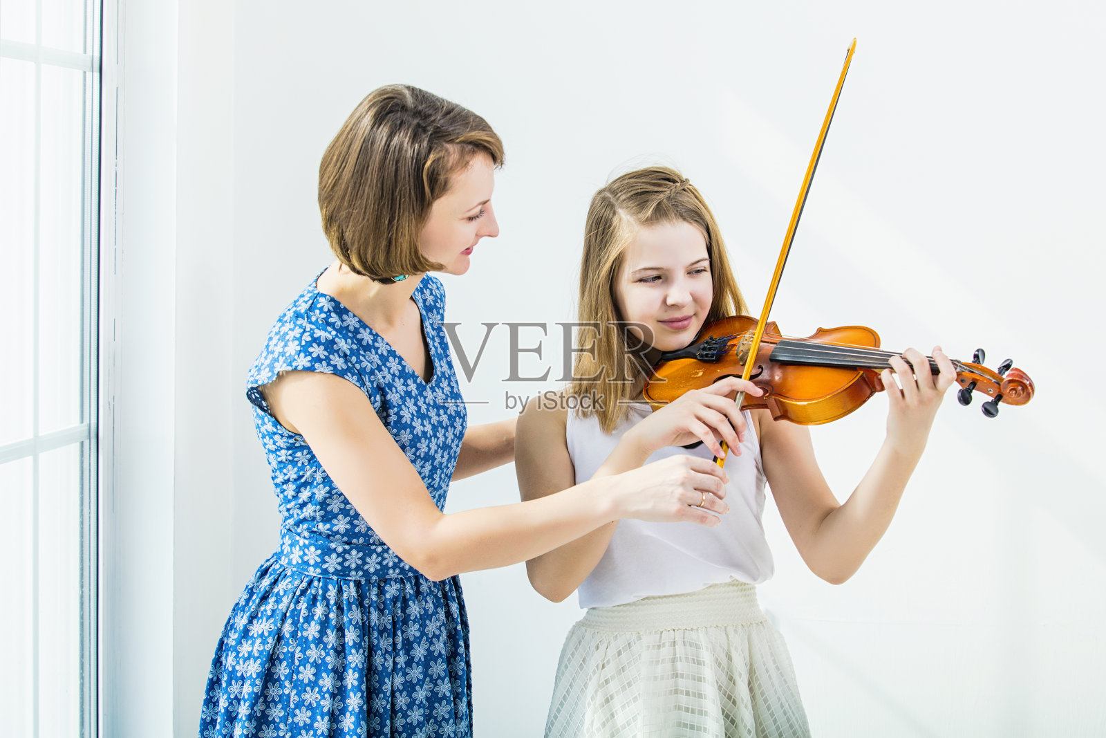 在有窗户的白色房间里，拉着小提琴的小女孩和老师在一起，美丽而快乐照片摄影图片