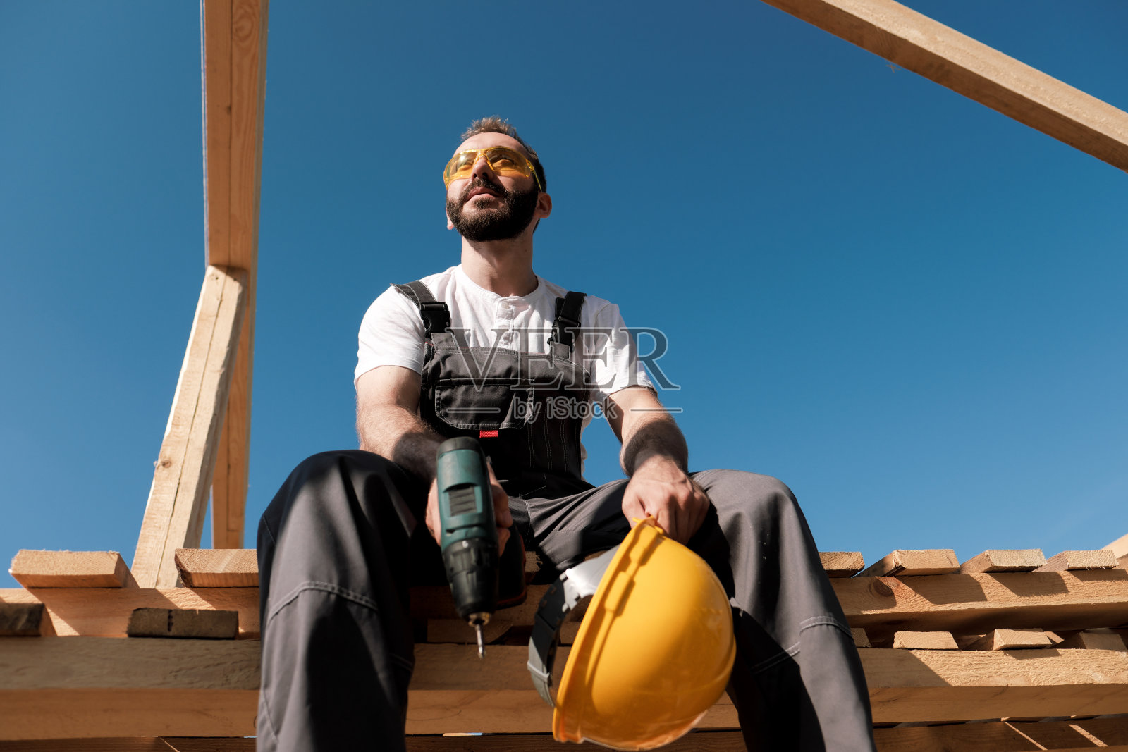建筑工人坐在木屋的屋顶边上，戴着黄色的头盔，穿着灰色的工作服。蓝天和晴朗的日子照片摄影图片