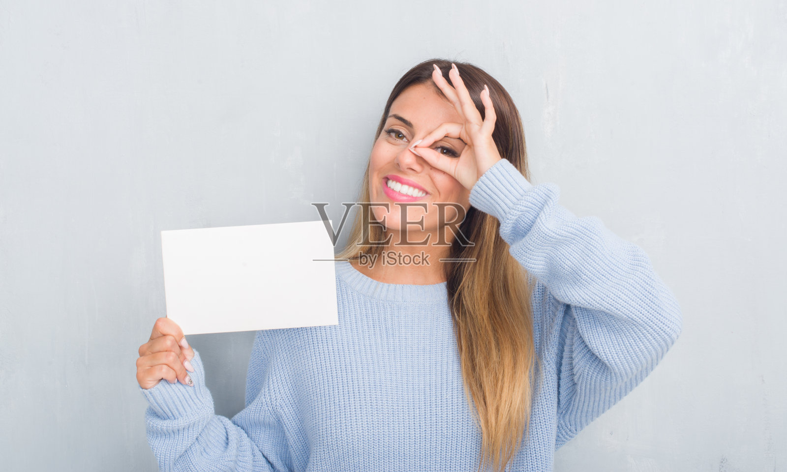 年轻的成年妇女在灰色的垃圾墙持有空白卡片与快乐的脸微笑做ok手势与手在眼睛透过手指看照片摄影图片