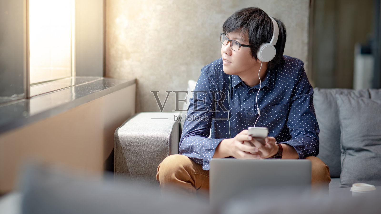 年轻的亚洲男子用智能手机在手机应用上听音乐，空闲时间坐在舒适的沙发上放松。生活空间中的城市生活方式。放松的概念照片摄影图片