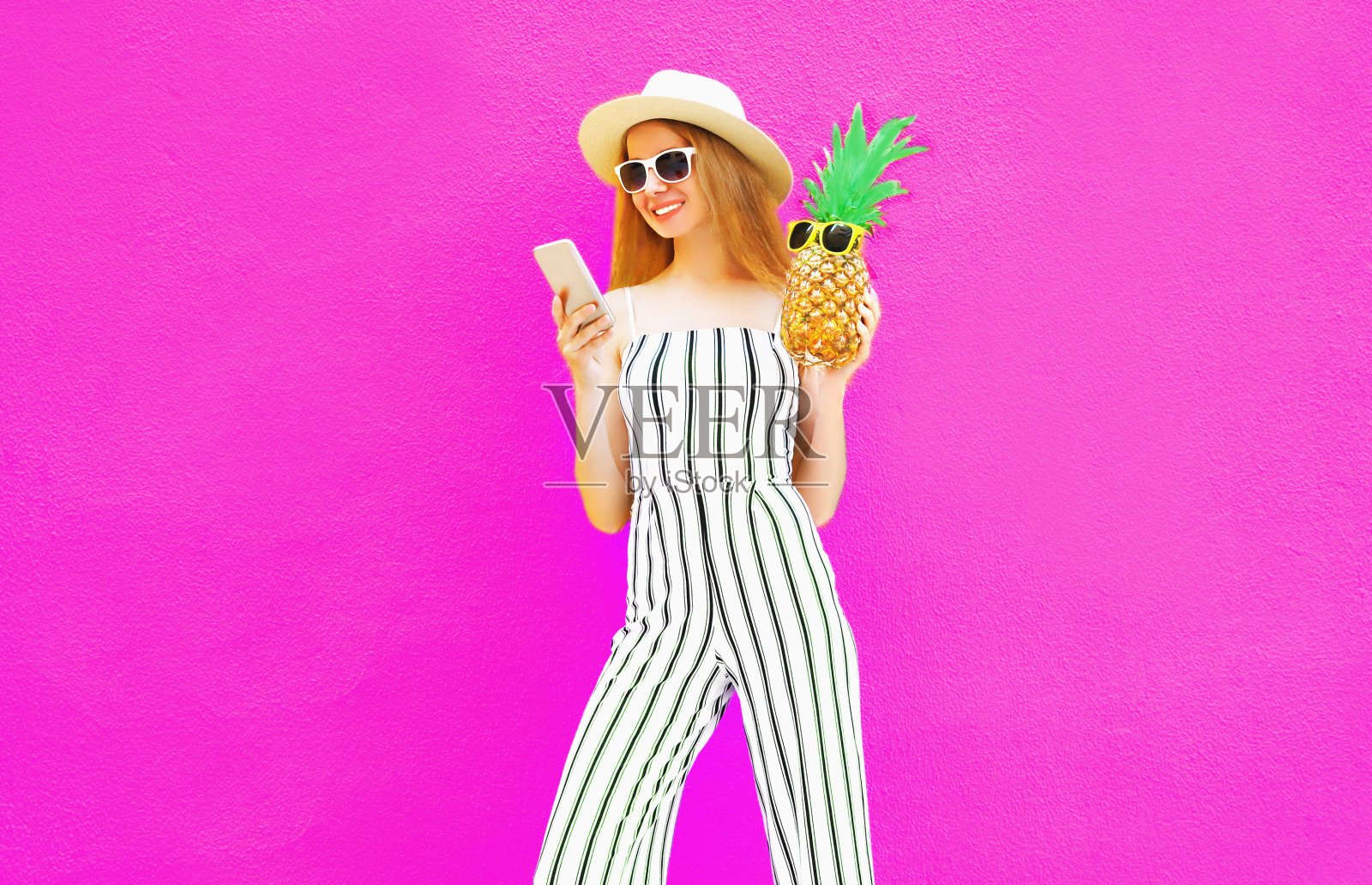 漂亮的女人拿着智能手机，菠萝在夏季圆帽，白色条纹连衣裤在彩色的粉红色墙壁背景照片摄影图片