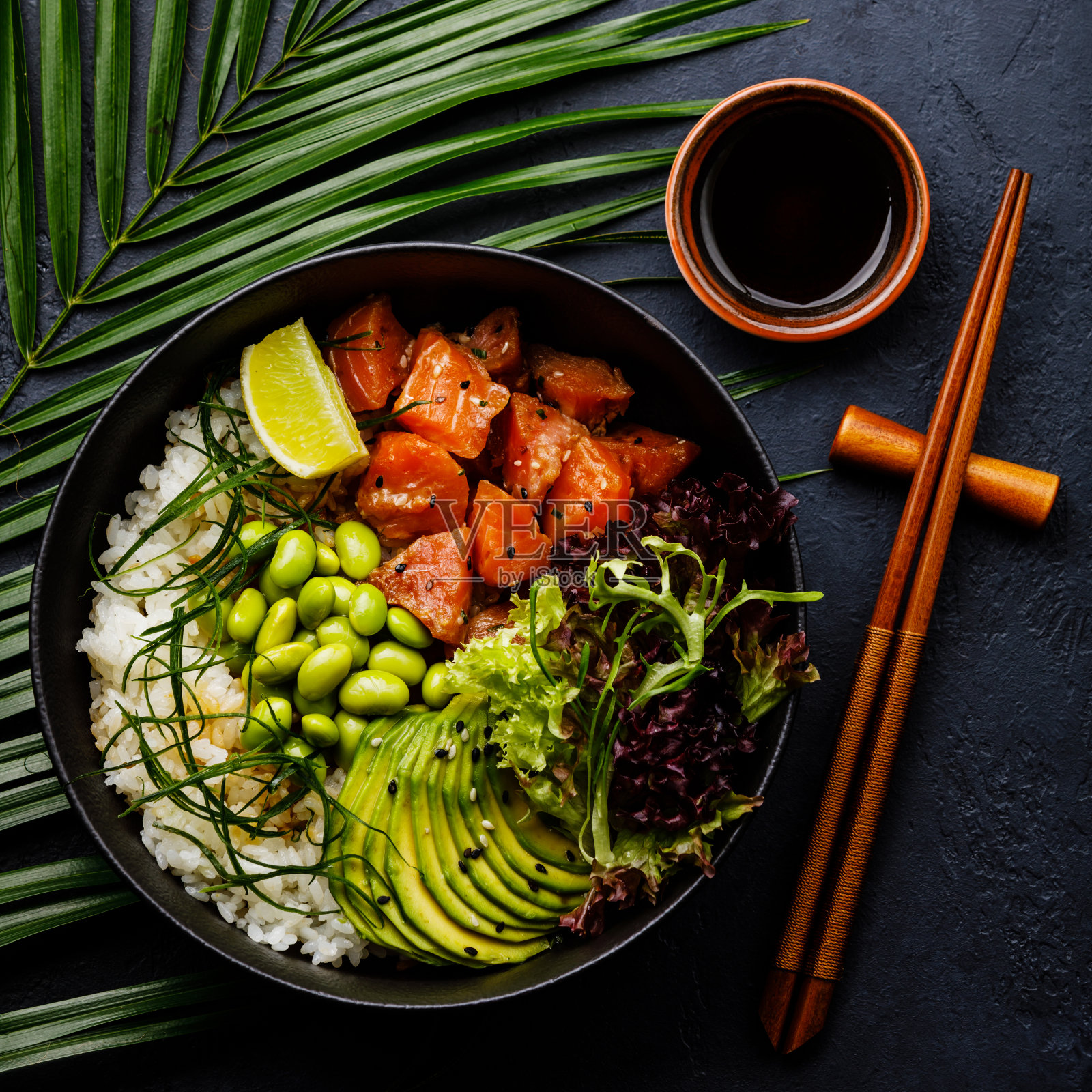 三文鱼戳碗生鱼沙拉亚洲流行食物，用大豆、毛豆、米饭、牛油果和莴苣放在碗里，在热带的叶子和深色的背景上照片摄影图片
