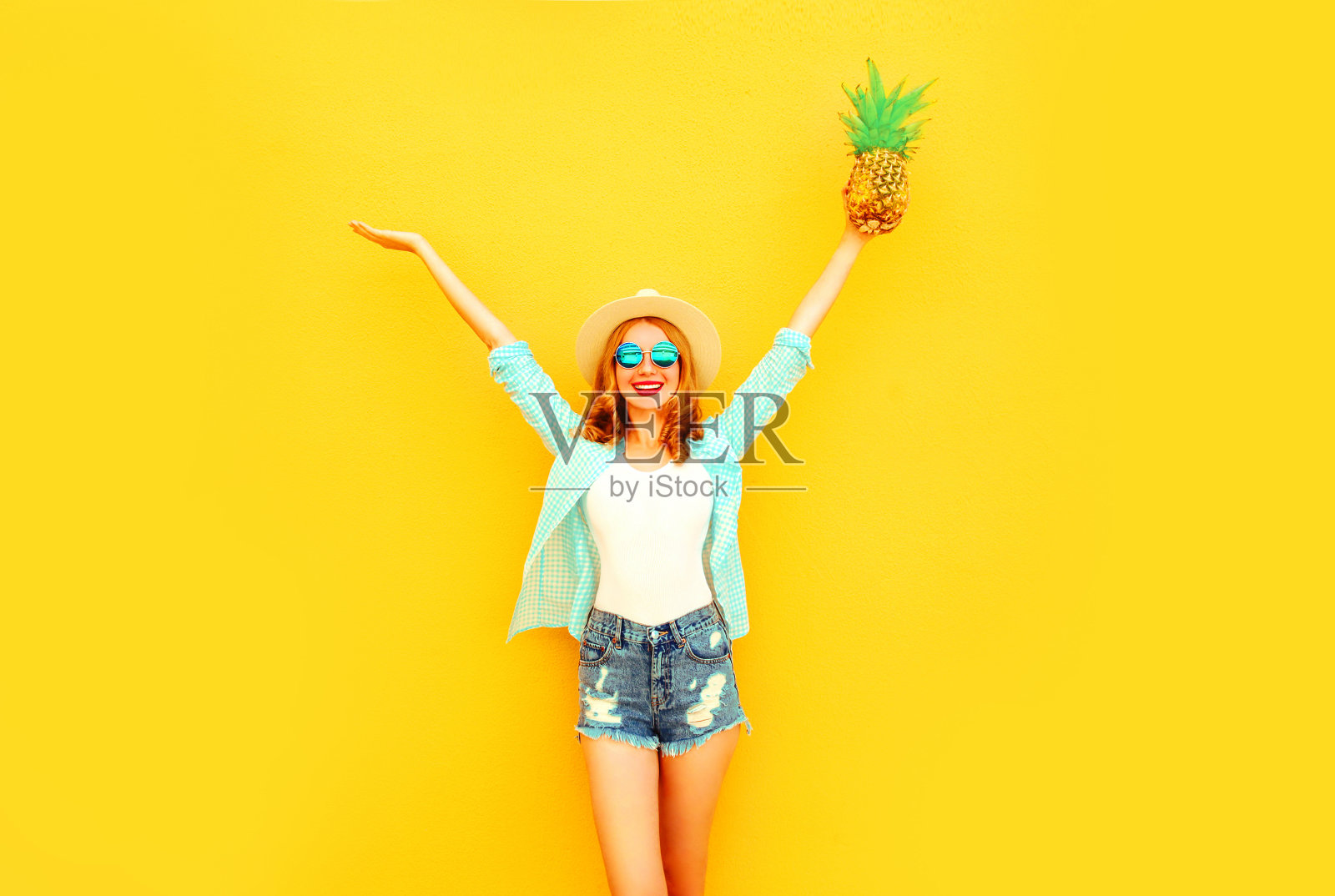 快乐微笑的年轻女子举起她的手与菠萝有乐趣的夏天草帽，太阳镜，短裤在彩色的黄色背景照片摄影图片