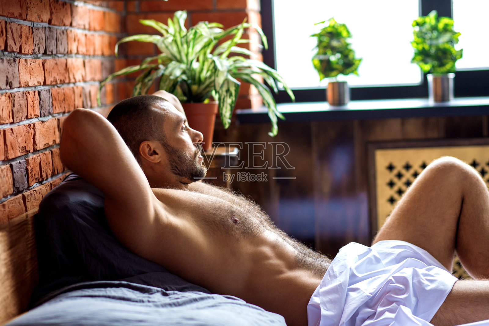 年轻的肌肉男模特躺在床上，看着窗户。照片摄影图片
