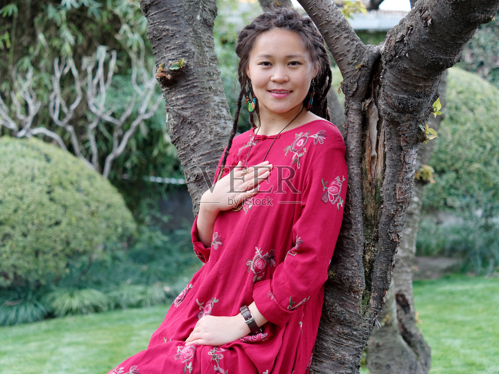 一个美丽的中国女孩的肖像，穿着红色长袍，梳着可爱的长发绺倚在树上，微笑着对着镜头，手放在她的胸口，善良友好的好女孩表现出善良。照片摄影图片