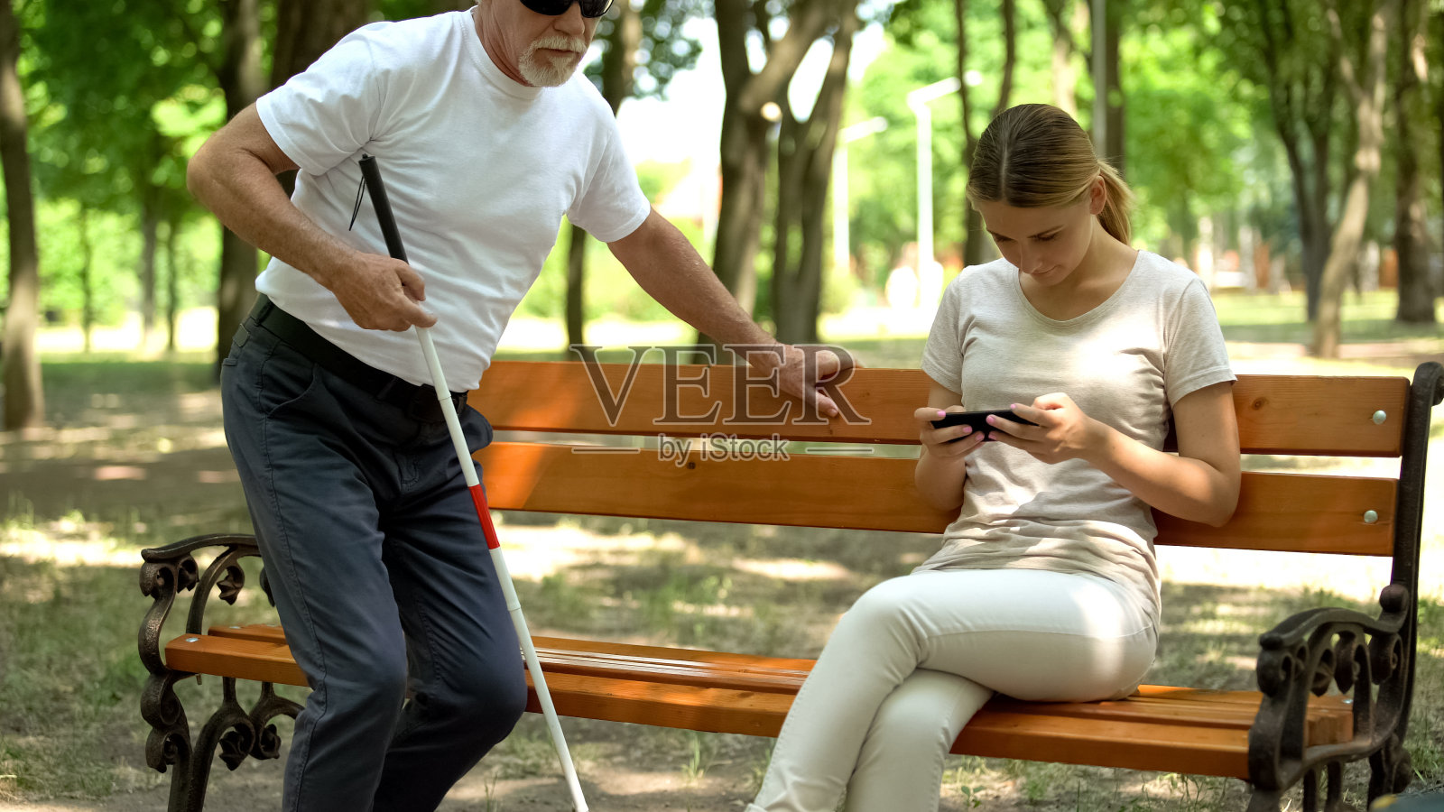 盲人试图坐在长椅上，年轻女士用手机发短信无视残疾人照片摄影图片
