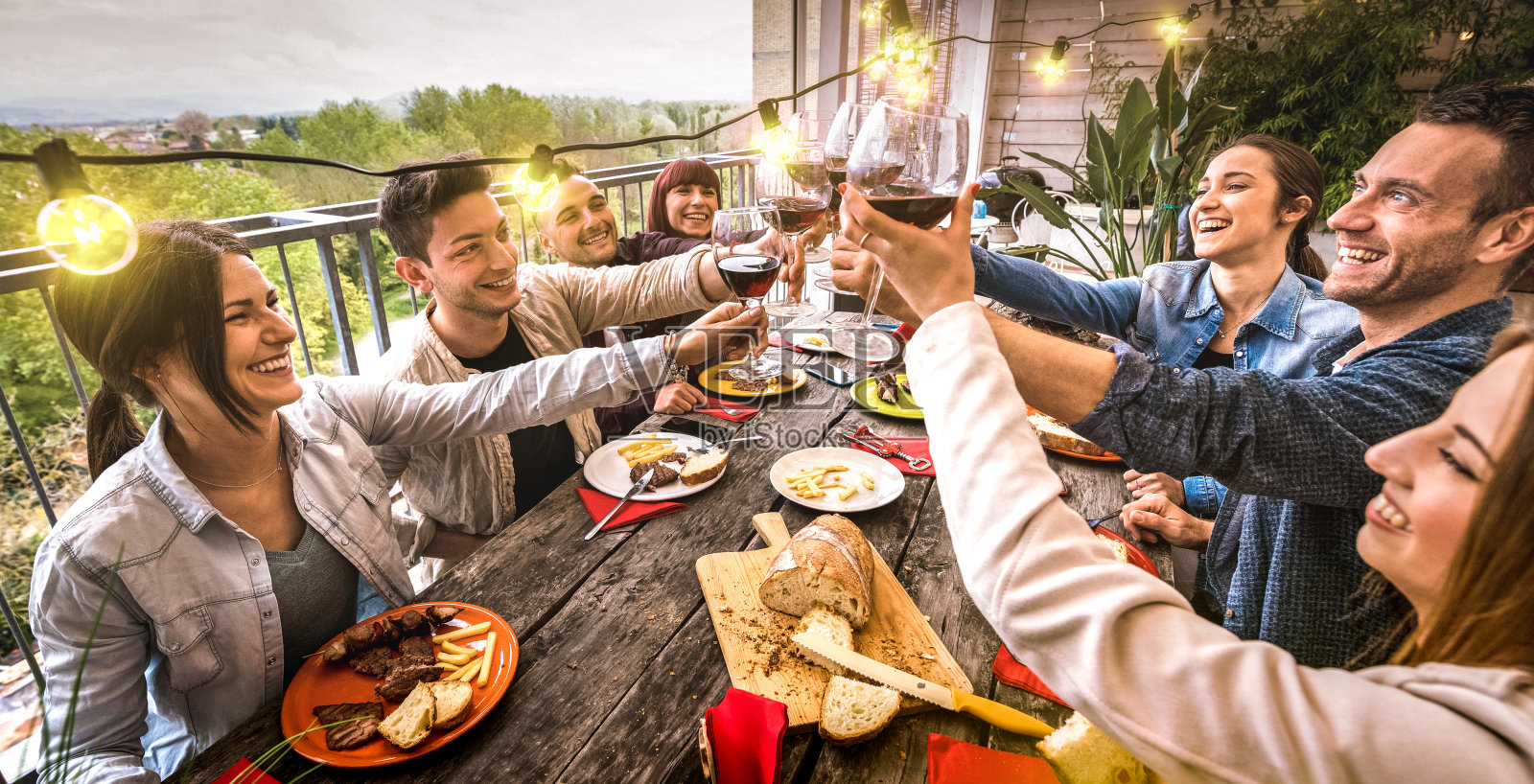 年轻人在户外别墅的晚餐聚会上一起举杯畅饮红酒-快乐的朋友在餐厅的露台上吃烧烤食物-温暖复古滤镜上的千禧生活方式概念-宽广的视野照片摄影图片