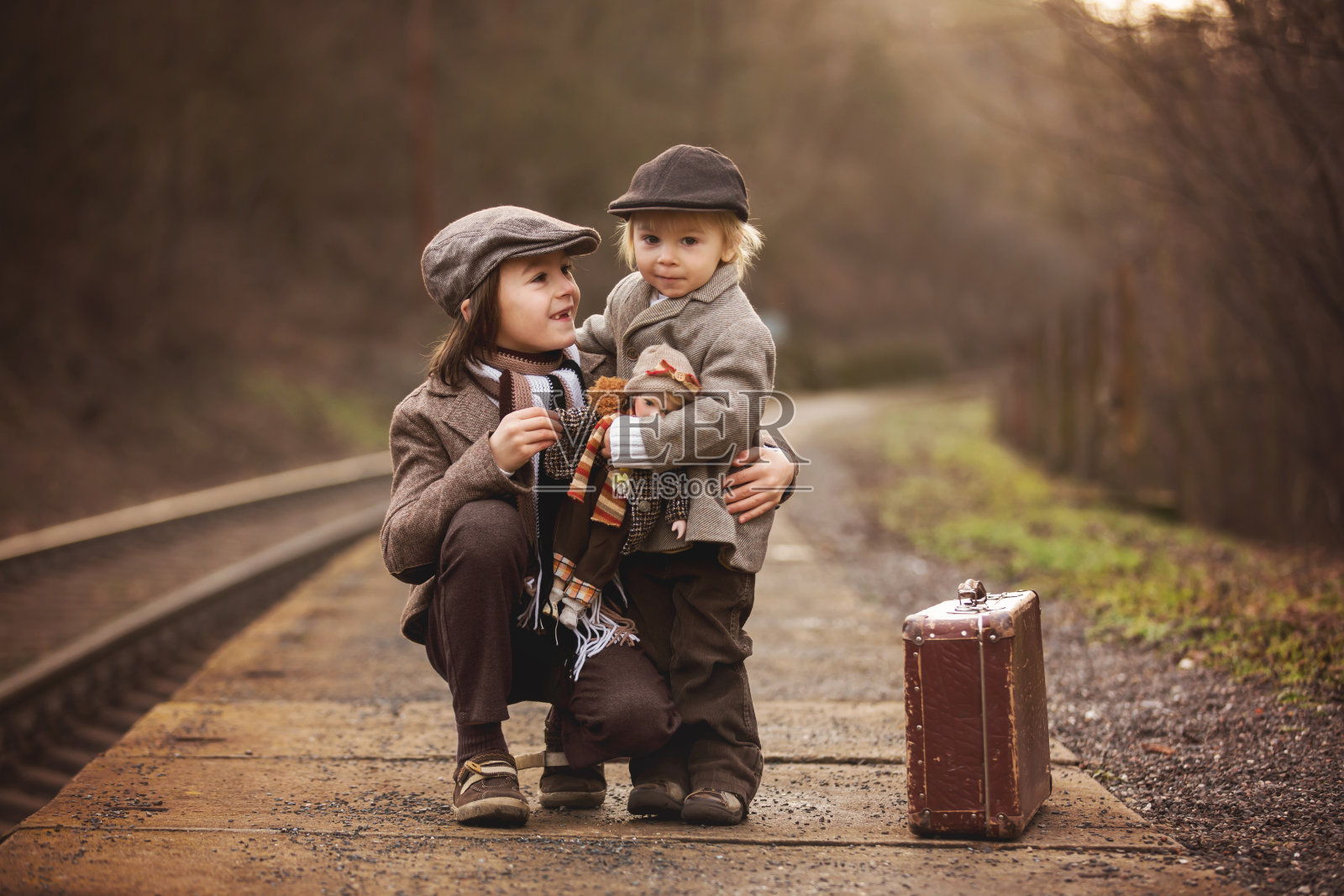 可爱的男孩在火车站，等待火车手提箱和漂亮的古董娃娃照片摄影图片