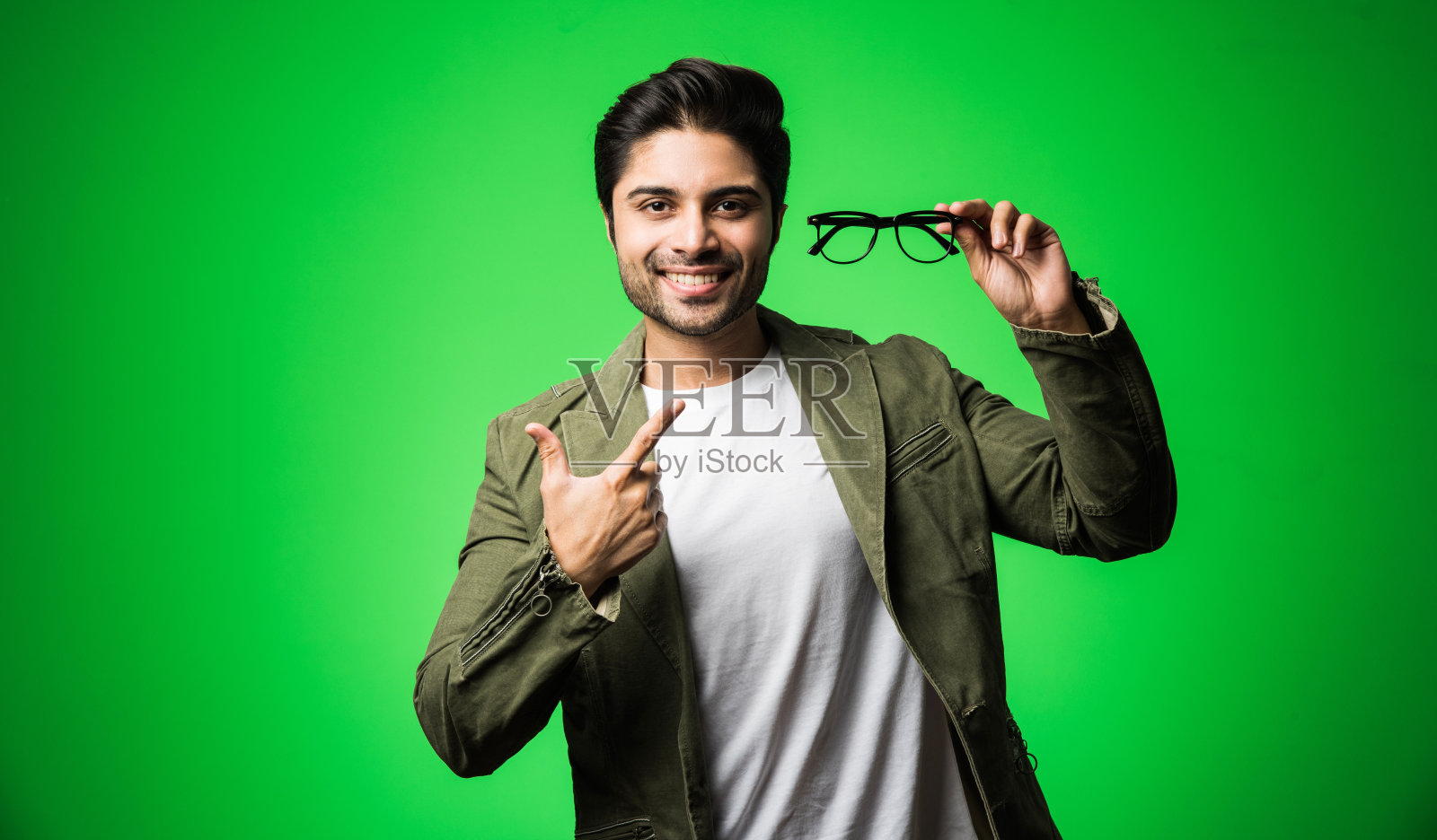 一名印度男子戴着眼镜，穿着t恤和夹克，孤立地站在绿色背景下照片摄影图片