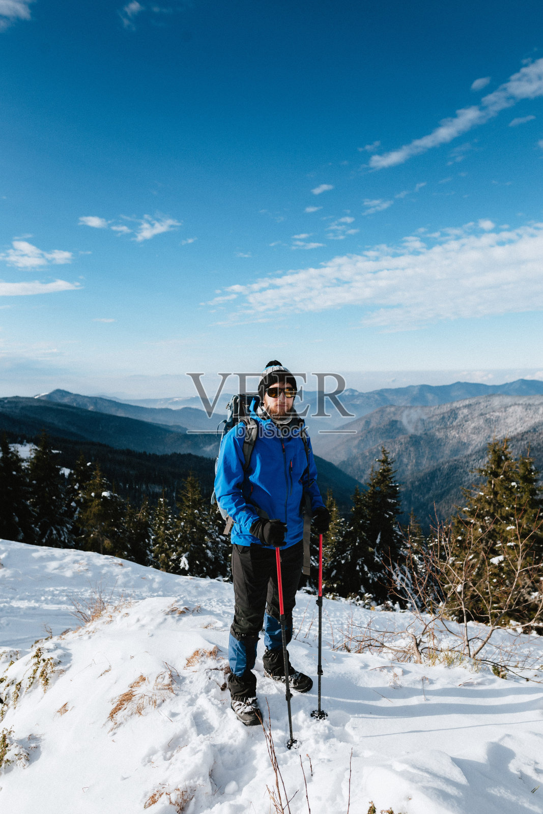 一名身穿蓝色夹克、手持登山杖的登山者，在天气好的冬季群山背景下，站着摆姿势照片摄影图片