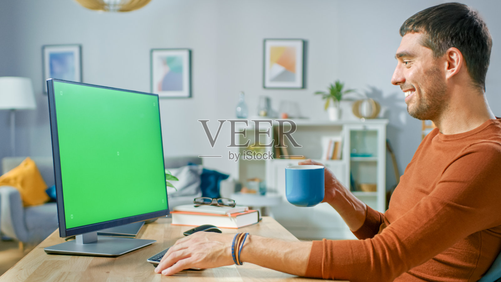 英俊的男人坐在他的书桌在家里使用个人电脑模拟绿色屏幕。他从杯子里喝饮料。照片摄影图片