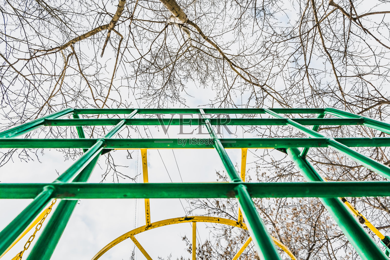 绿色和黄色的金属攀爬网格与蓝天白云，白杨树和枫树的树枝在操场上照片摄影图片