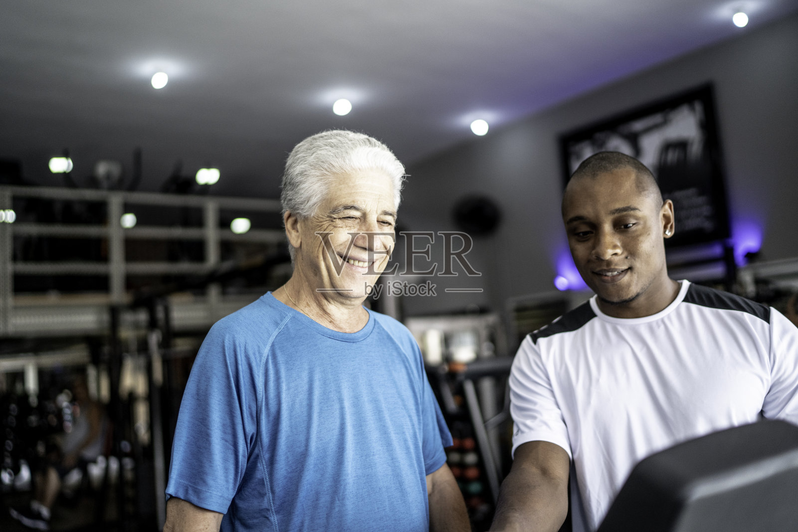 一位老人在私人教练的帮助下在健身房的跑步机上慢跑照片摄影图片