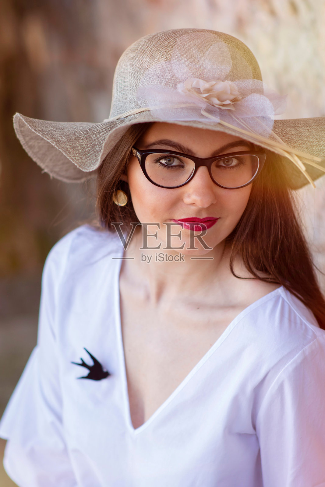 一位戴着眼镜和帽子、面带微笑的年轻女子站在一堵旧砖石墙前。照片摄影图片