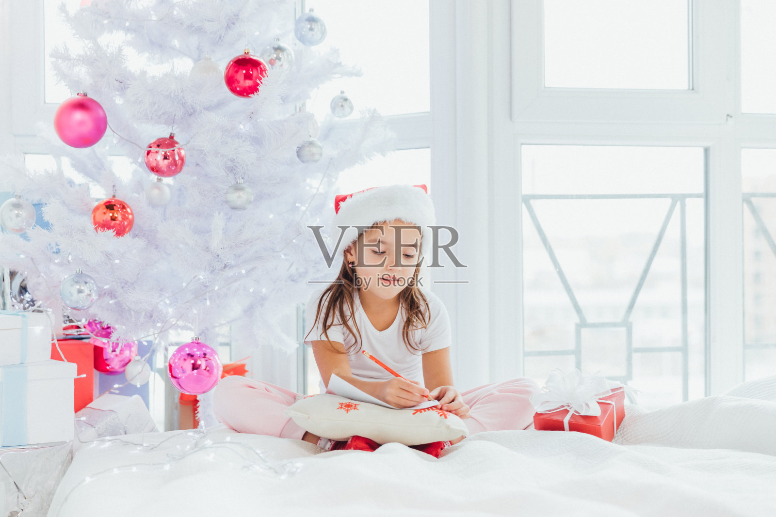 在寒冷的冬天的早晨，小女孩戴着圣诞老人的帽子在圣诞树旁给圣诞老人写信。照片摄影图片