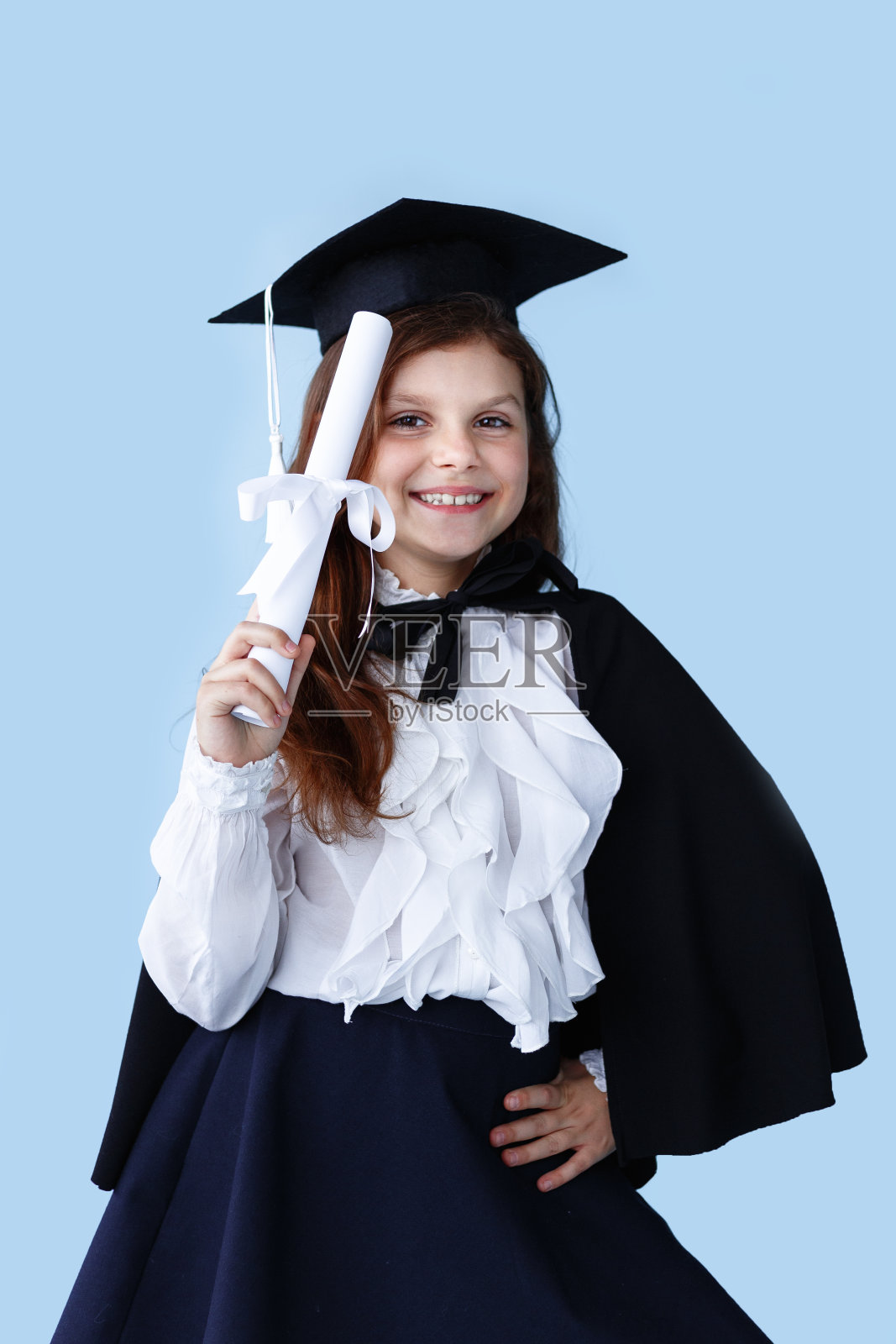 一个快乐的小女孩戴着毕业帽，站在蓝色背景上，展示着一个成功的时刻。照片摄影图片