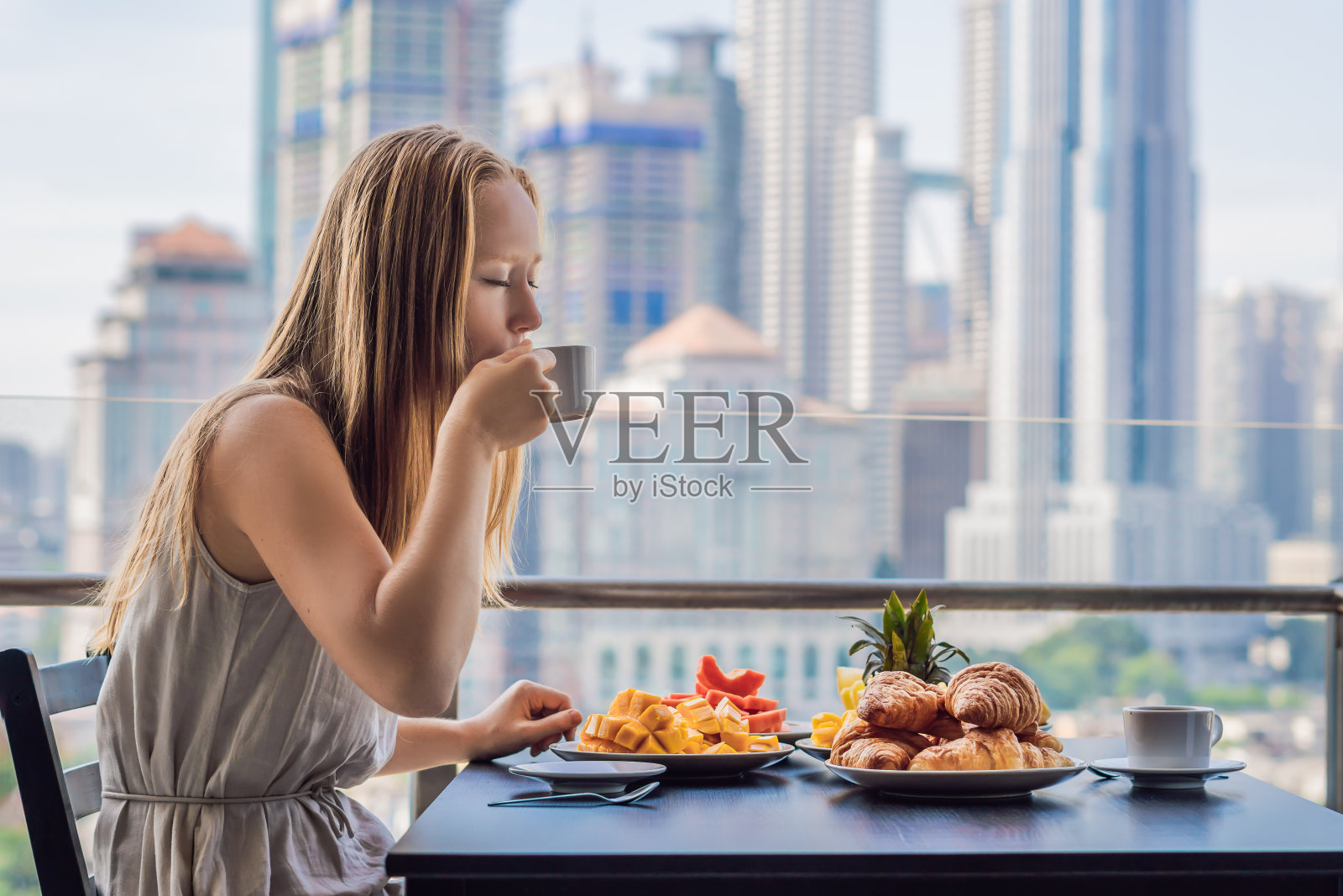 一位年轻女子正在阳台上吃早饭。阳台上的早餐桌上放着咖啡、水果和牛角面包，背景是大城市照片摄影图片