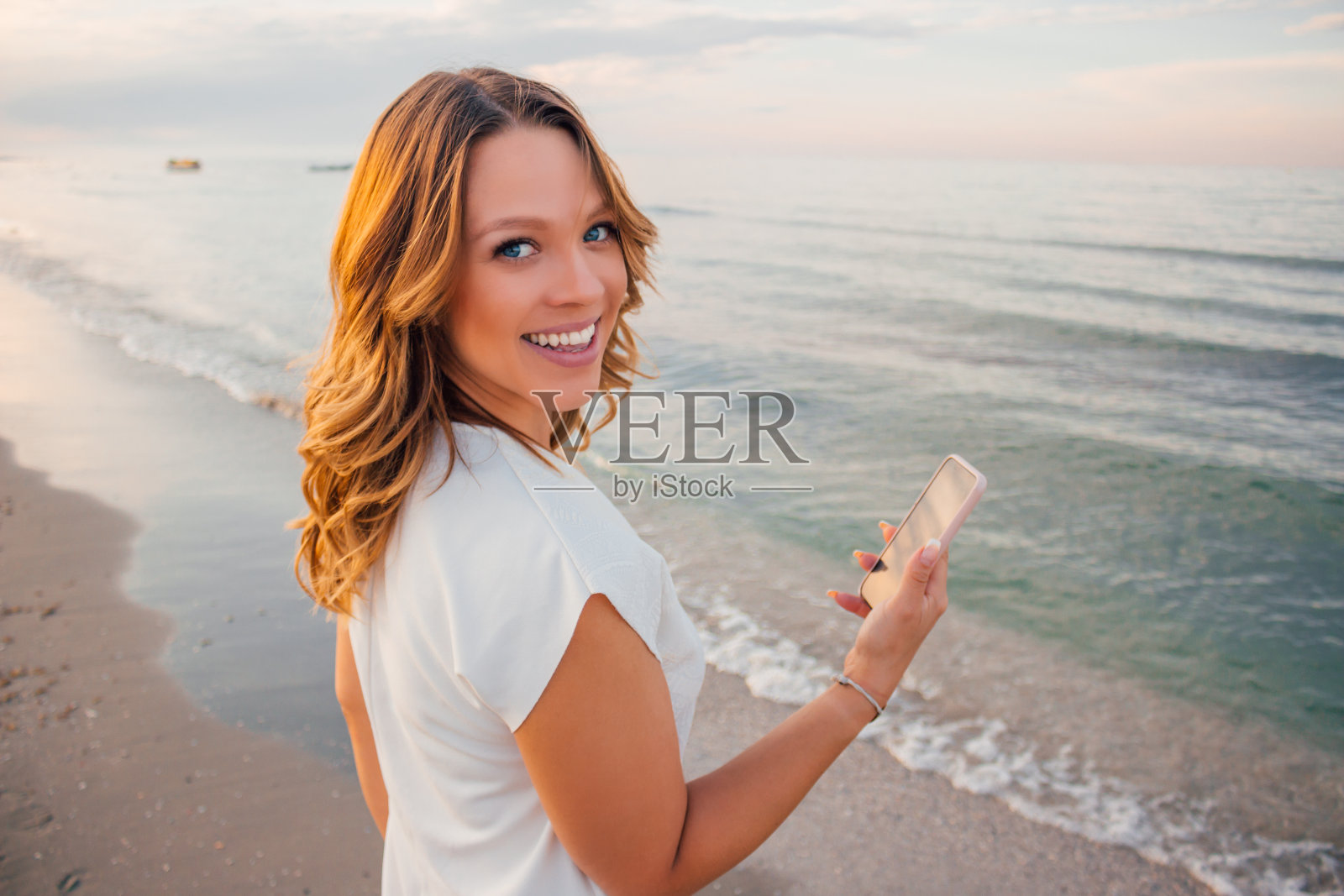 年轻美丽的微笑金发曲线模特在海滩上摆姿势与智能手机在她的手穿着白色连衣裙-加大尺寸的长卷发模特在海边看相机而发短信照片摄影图片