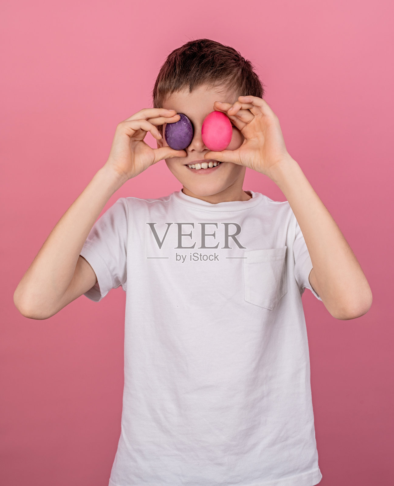 微笑的男孩用五颜六色的鸡蛋遮盖眼睛，孤立在粉红色的背景照片摄影图片