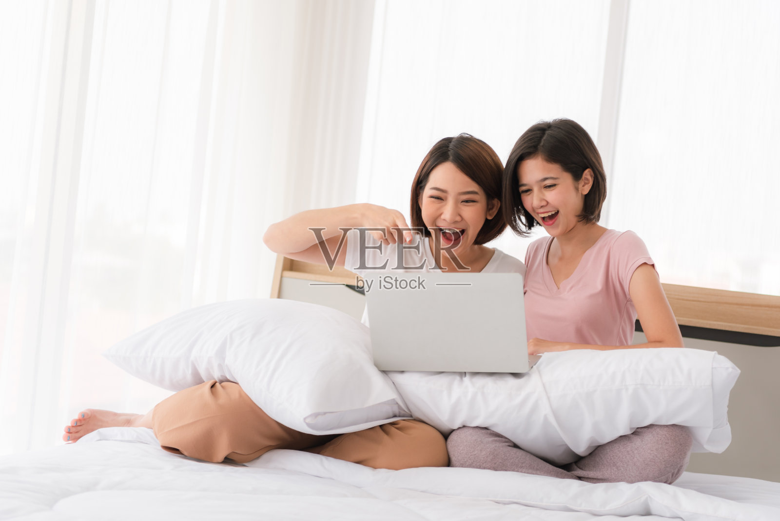 快乐随意的两个漂亮女人坐在家里的床上用笔记本电脑工作。照片摄影图片
