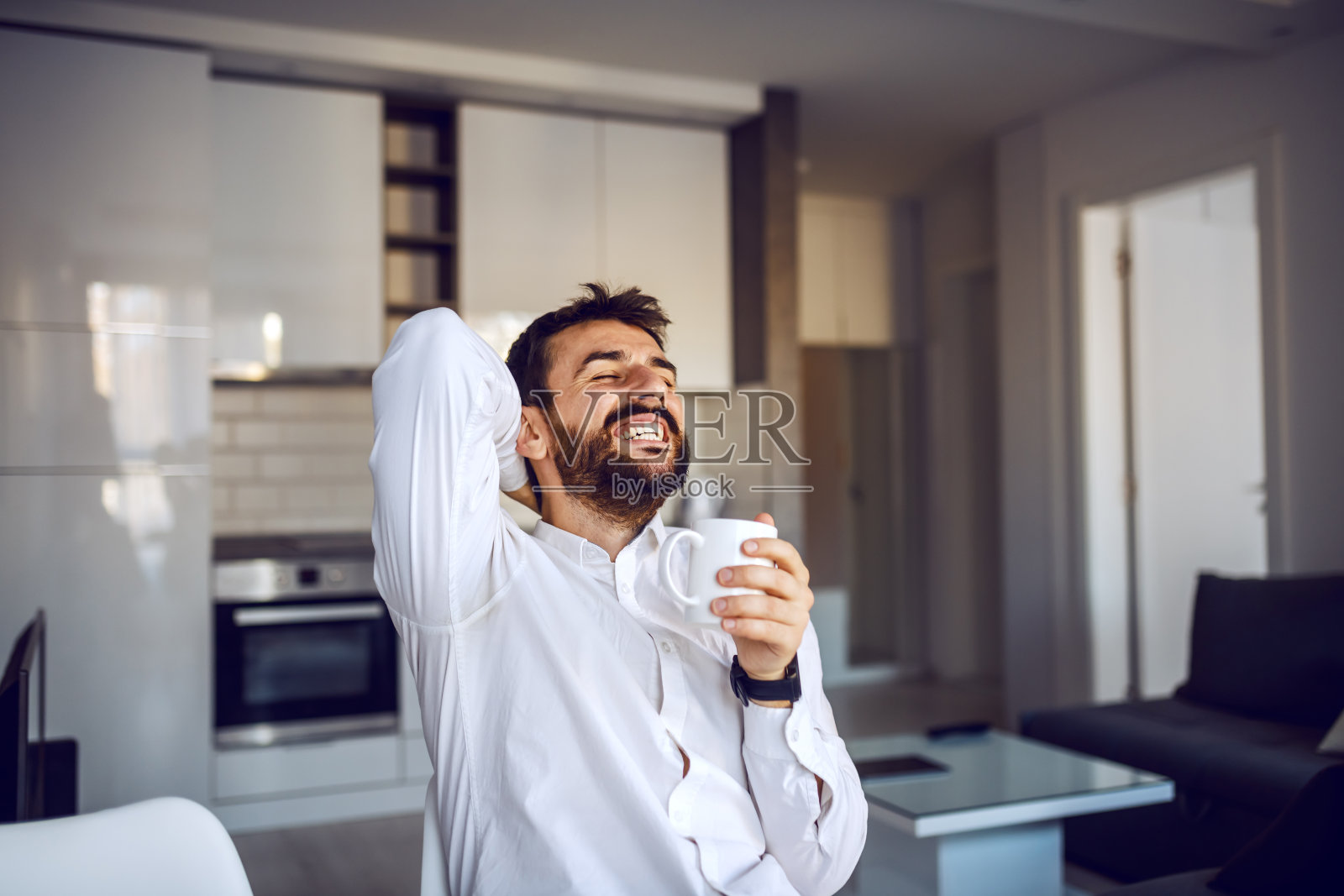 年轻微笑有吸引力的胡子男子坐在家里拿着杯新鲜的早晨咖啡和放松。照片摄影图片