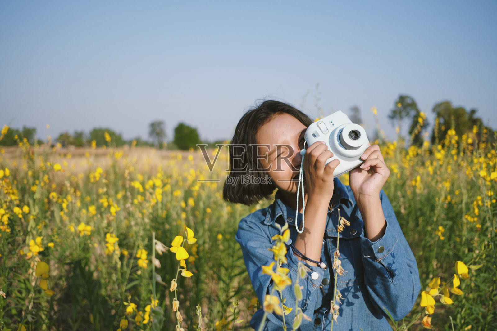可爱的快乐亚洲年轻女孩使用相机穿牛仔裤在美丽的黄花花园。照片摄影图片