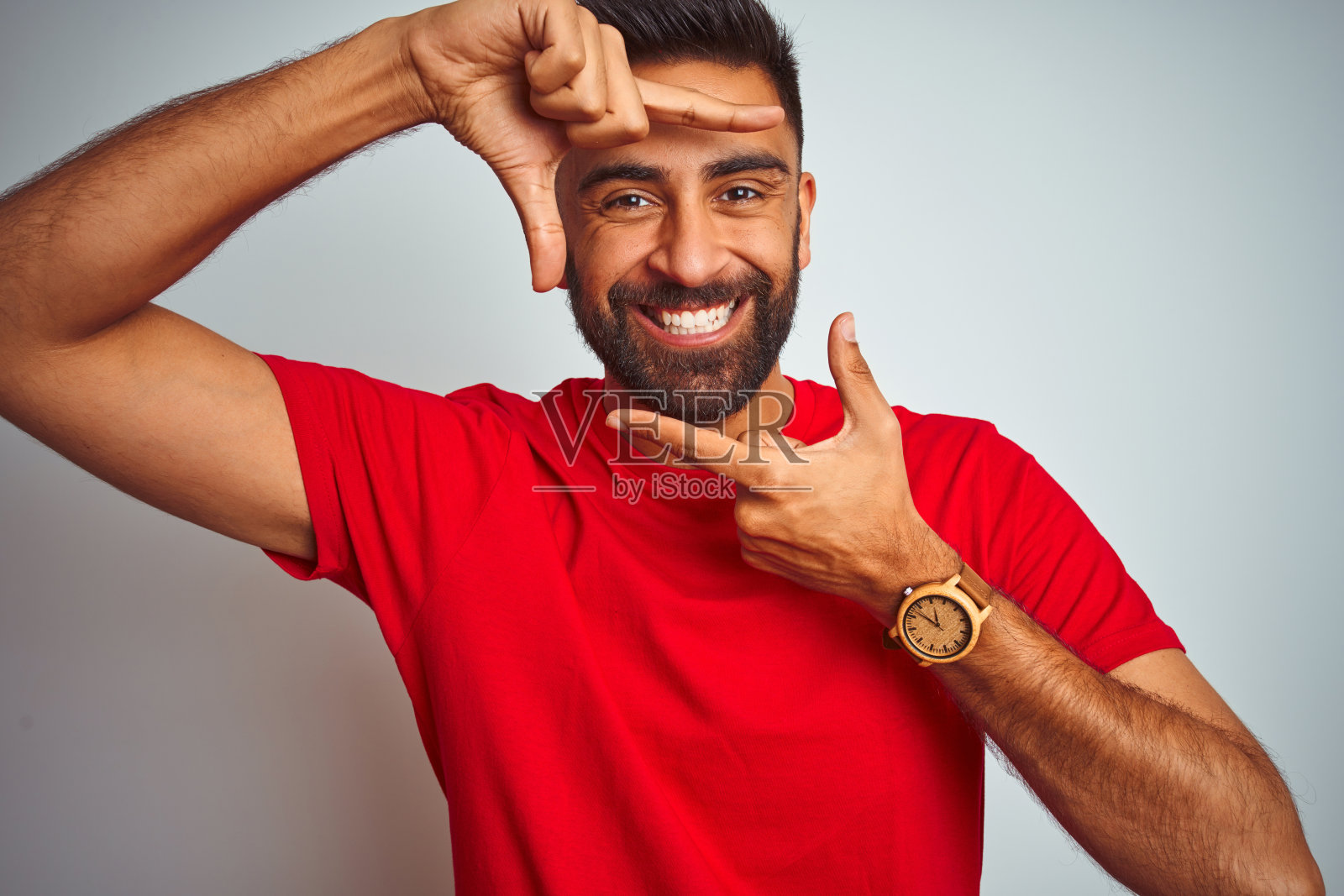 年轻的印度男子穿着红色t恤，在孤立的白色背景上微笑，用手和手指做框架，快乐的脸。创意和摄影概念。照片摄影图片