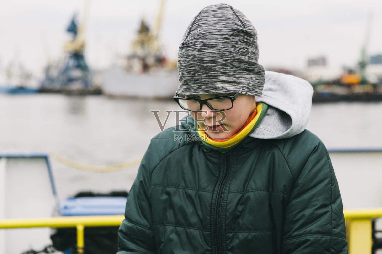 十几岁的男孩戴着冬天的帽子、外套和眼镜认真地向下看，而站在港口的码头旁边。照片摄影图片