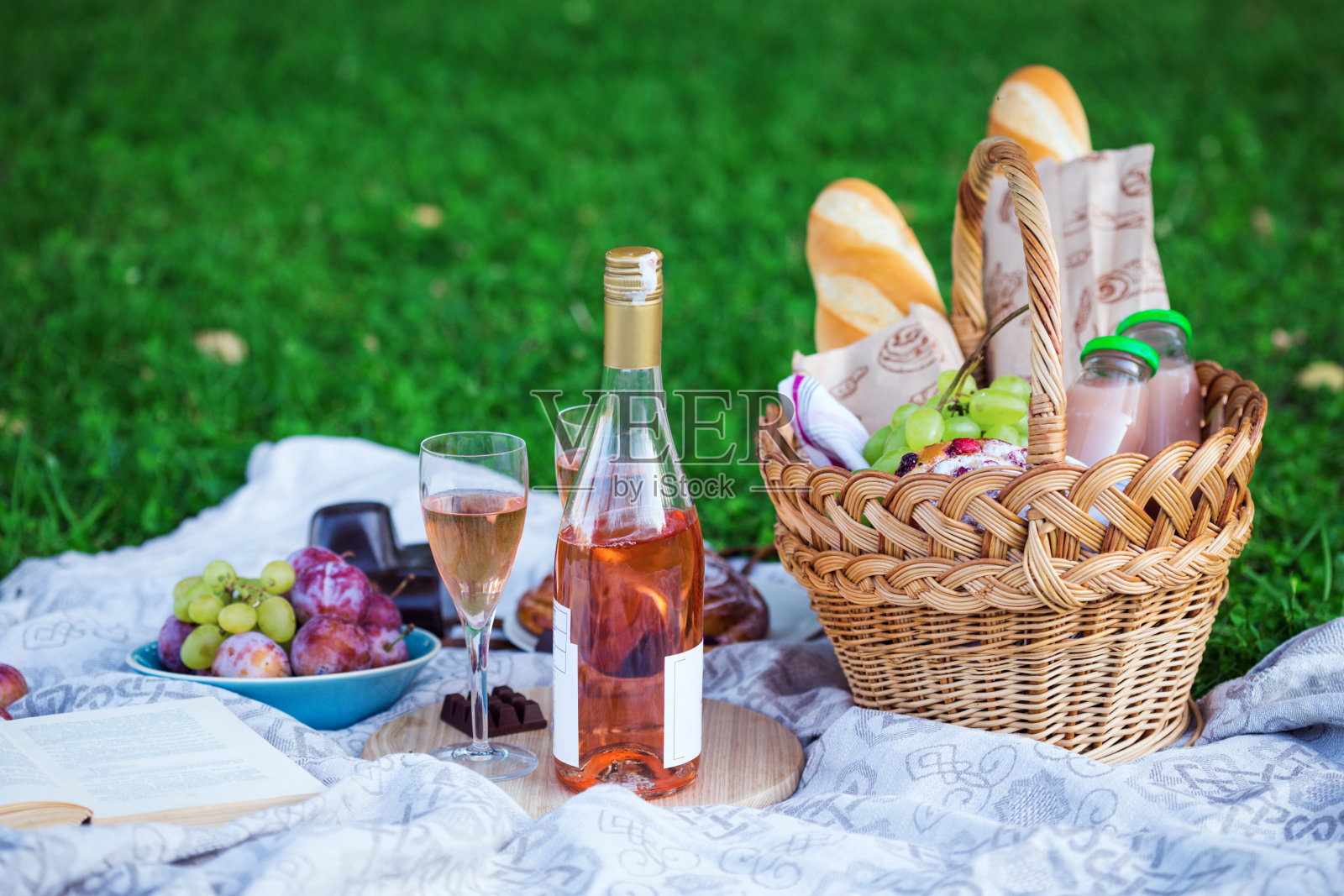 夏天在草地上野餐。长棍面包、葡萄酒、玻璃杯、葡萄和面包卷装在篮子里，放在绿草地上照片摄影图片