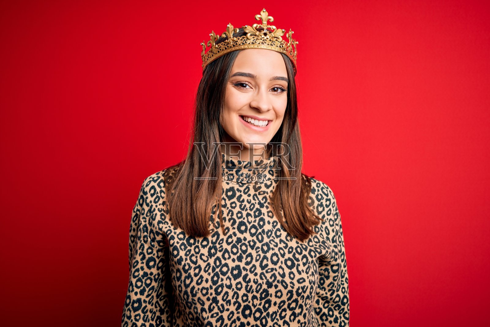 年轻美丽的黑发女子戴着金色皇冠皇后在孤立的红色背景与快乐和凉爽的微笑在脸上。幸运的人。照片摄影图片