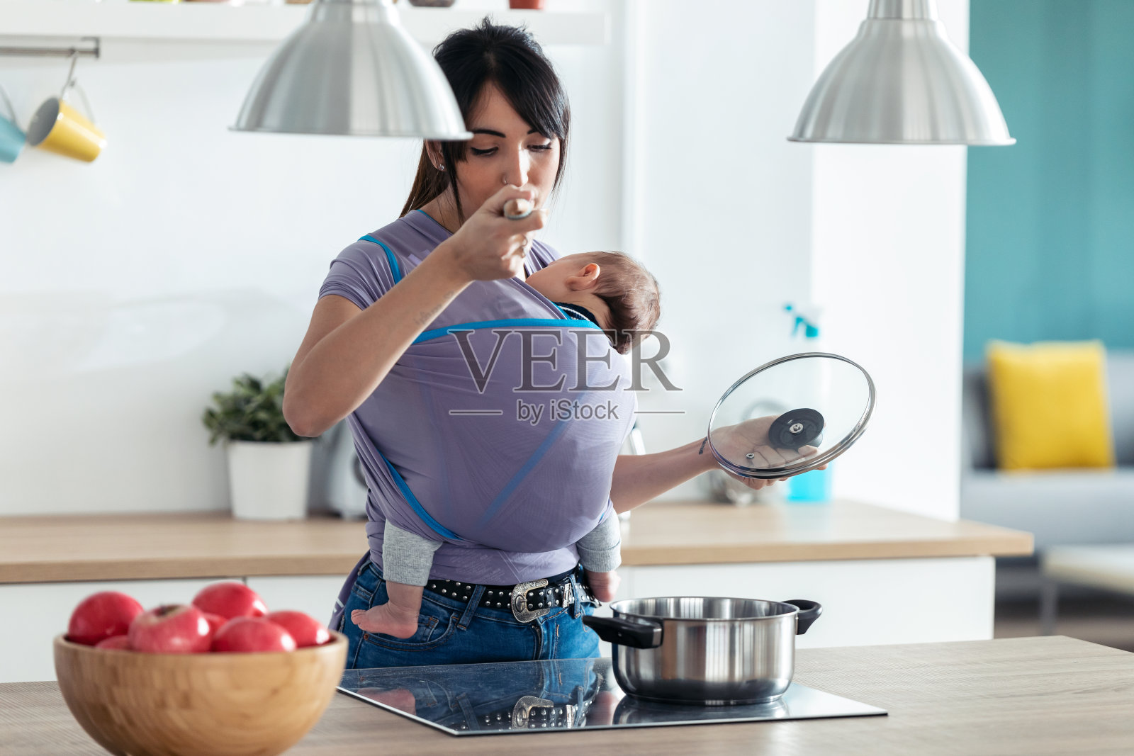 年轻漂亮的妈妈带着小宝宝在厨房里一边做饭一边品尝食物。照片摄影图片