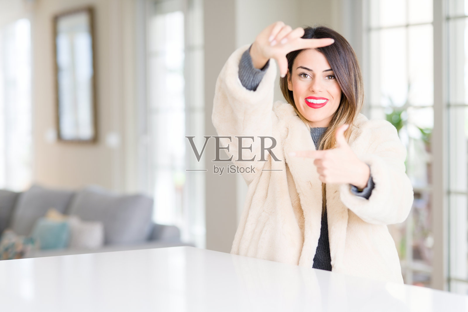 年轻美丽的女子穿着冬季皮大衣在家里微笑做框架与手和手指与幸福的脸。创意和摄影概念。照片摄影图片