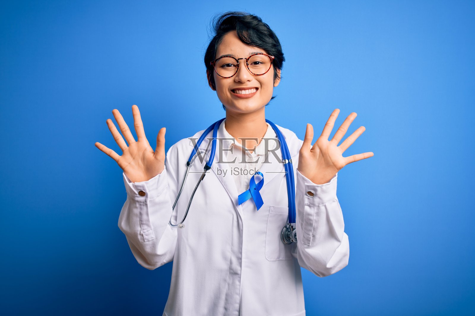 年轻美丽的亚洲医生女孩穿着听诊器和外套与蓝色癌症丝带显示和指着10号手指，同时自信和快乐的微笑。照片摄影图片