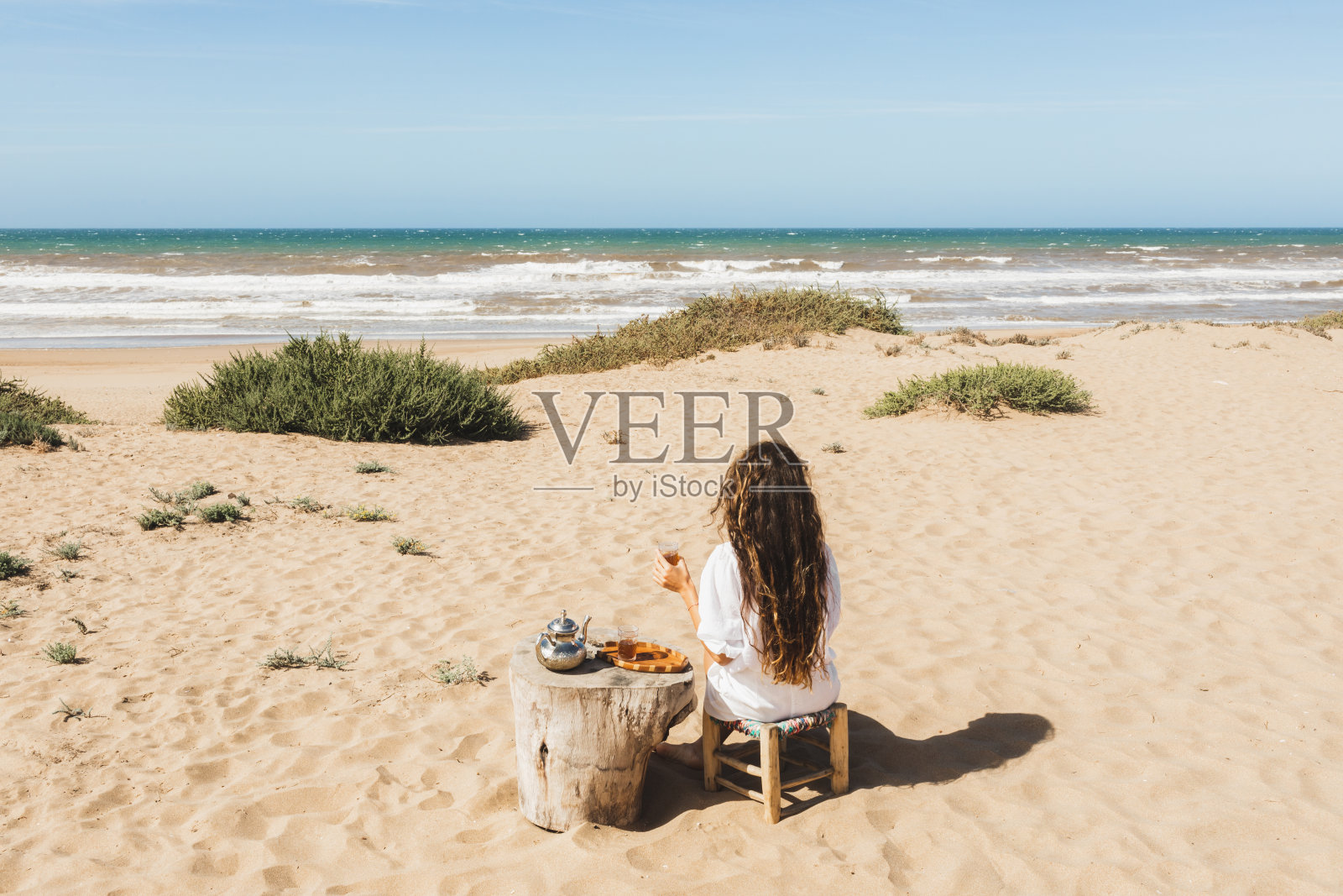在摩洛哥的海洋沙滩上，一名妇女在享受传统的摩洛哥薄荷茶。视图从后面。照片摄影图片