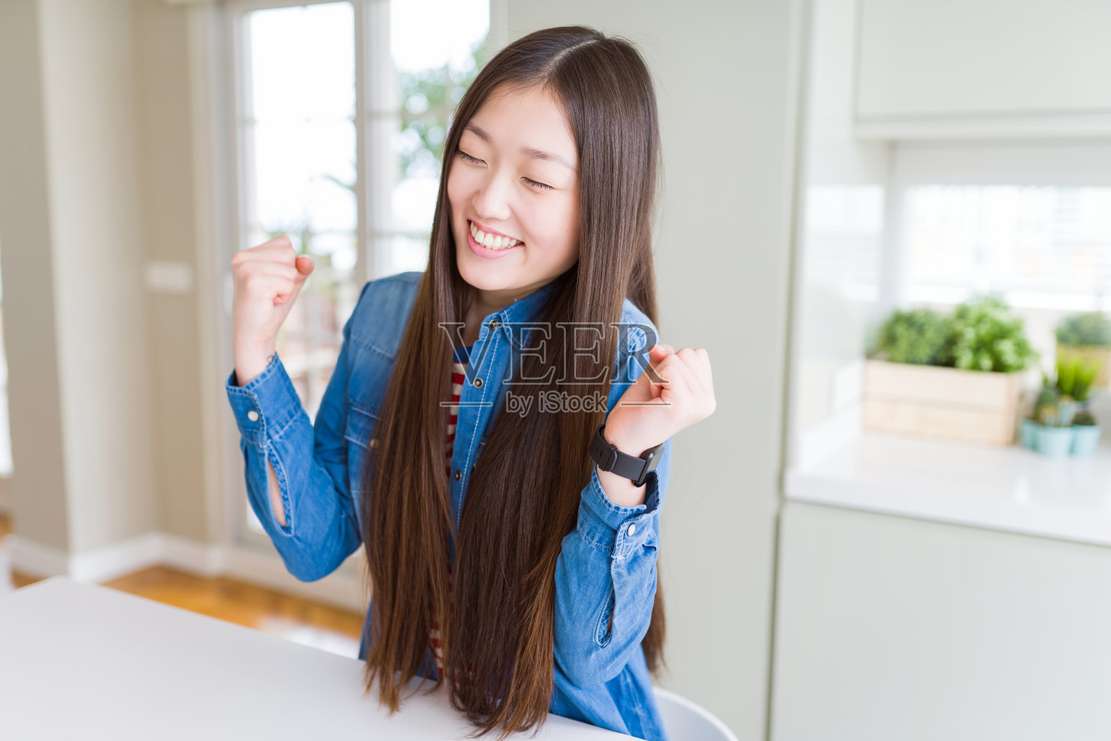 美丽的亚洲女人穿着牛仔夹克在白色的桌子兴奋的成功与双臂举起庆祝胜利微笑。赢家的概念。照片摄影图片