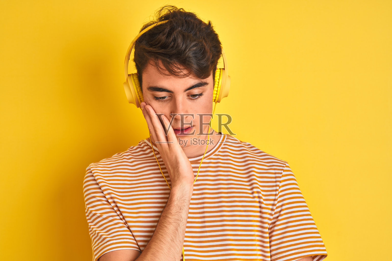 十几岁的男孩戴着耳机在孤立的黄色背景下思考，看起来疲倦和厌倦的抑郁症问题交叉双臂。照片摄影图片