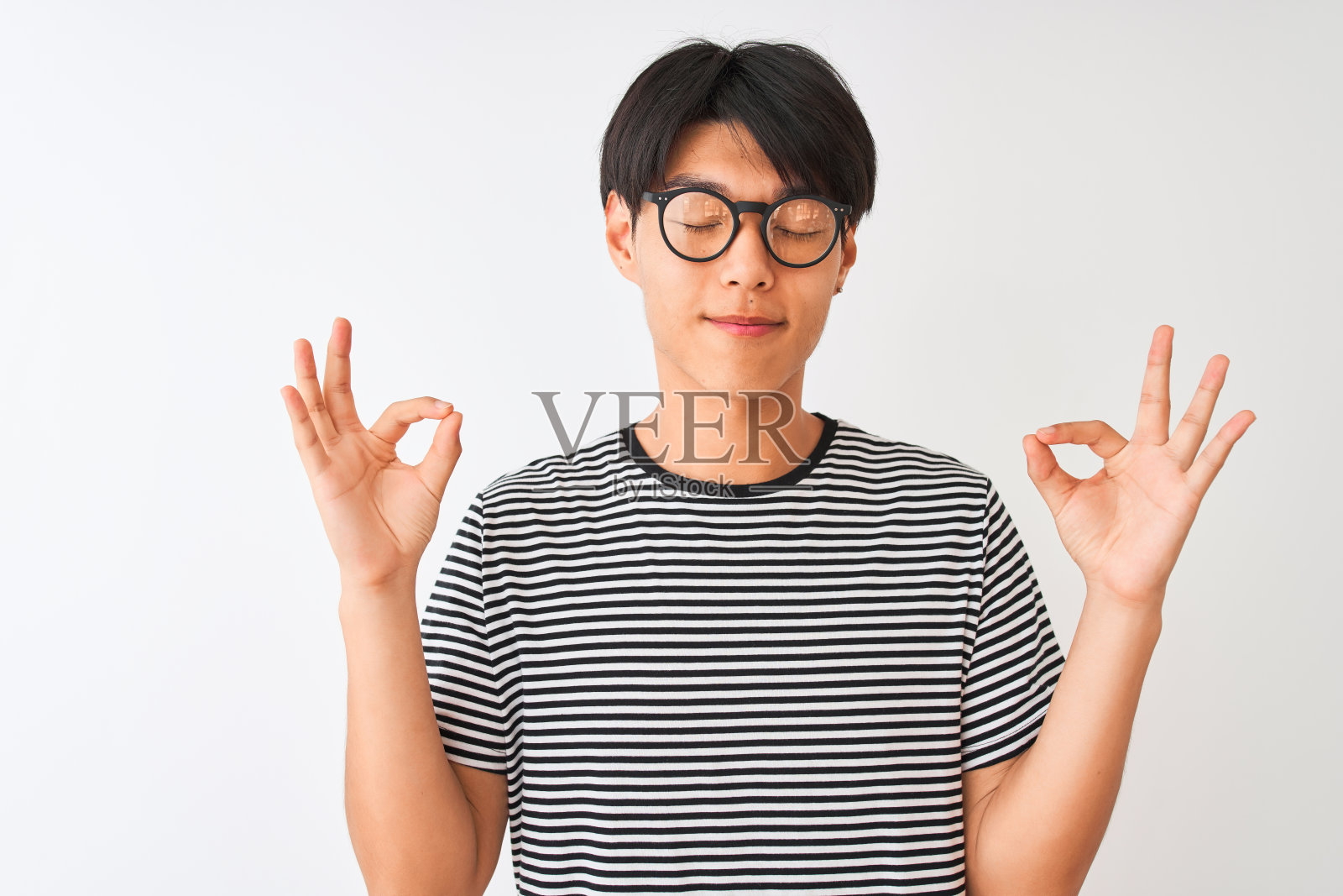 一名中国男子戴着眼镜，身穿海军蓝条纹t恤，站在孤立的白色背景下，闭着眼睛微笑着，用手指做冥想手势。瑜伽的概念。照片摄影图片