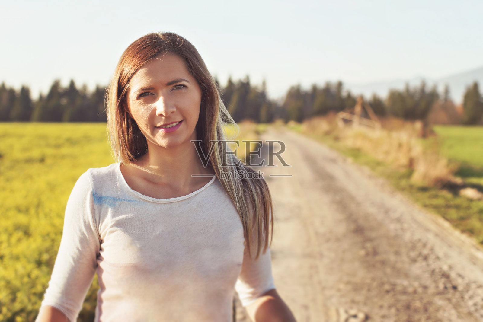 一名年轻女子站在尘土飞扬的乡村道路上，被午后的阳光照亮，背景是模糊的田野和森林，照片色调温暖照片摄影图片