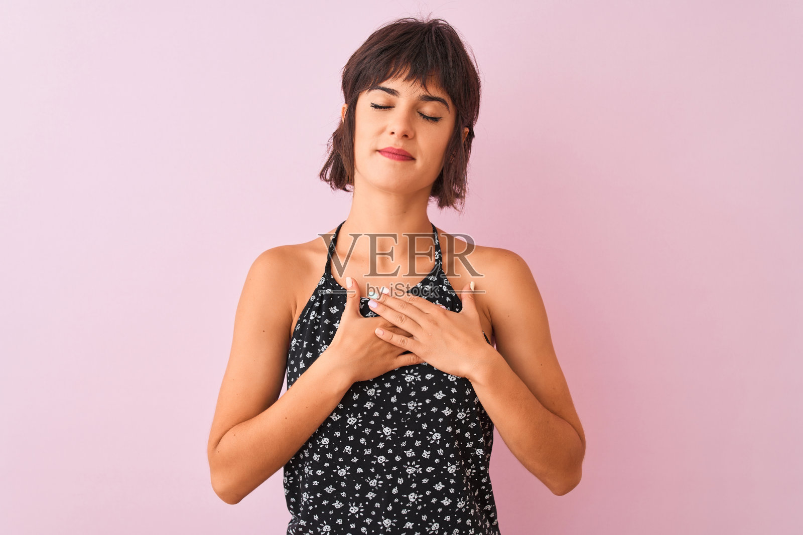 年轻美丽的女人穿着黑色碎花连衣裙站在孤立的粉红色背景微笑与手在胸前闭上眼睛和感激的手势在脸上。健康的概念。照片摄影图片