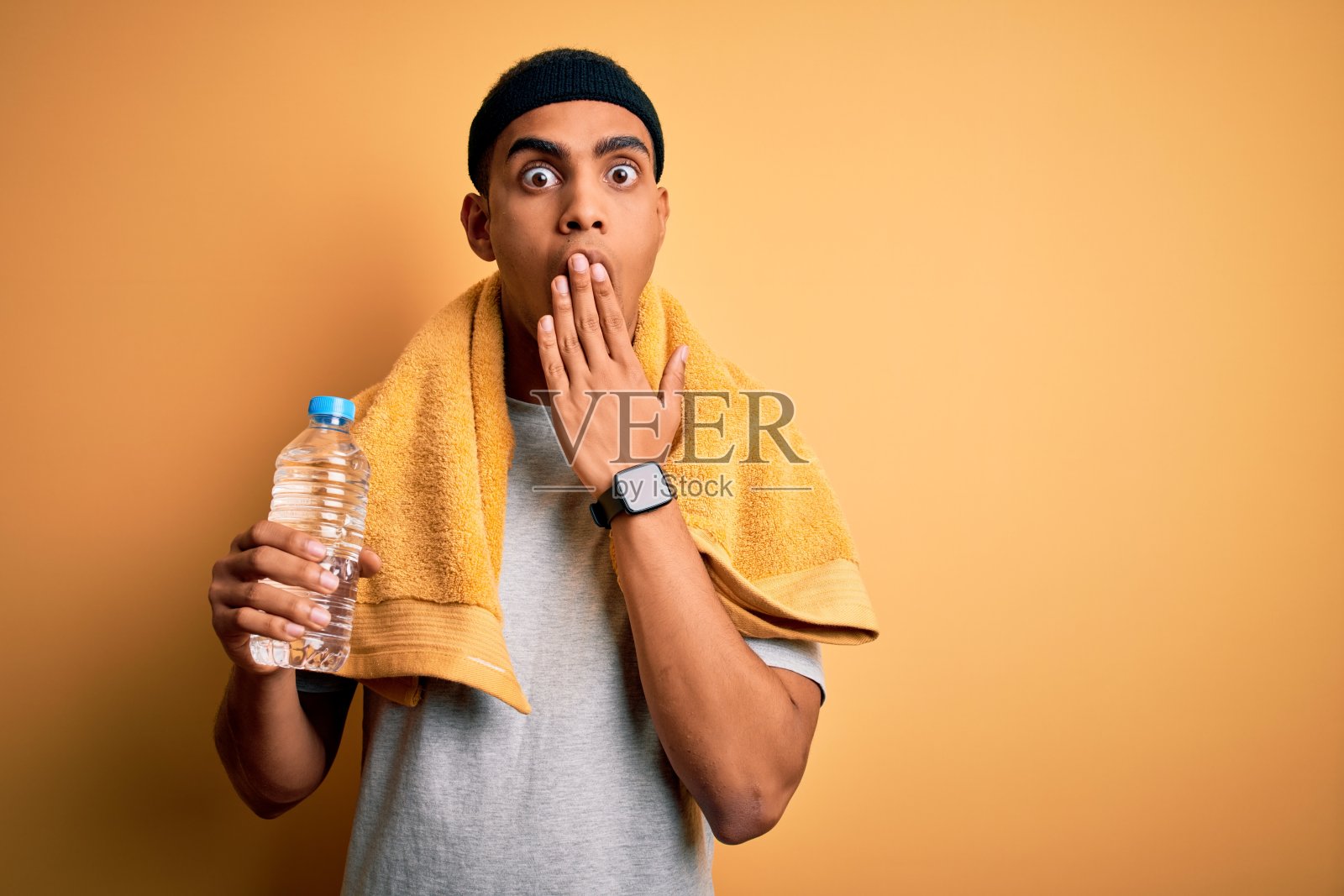 年轻英俊的非裔美国运动员戴着毛巾，喝着水，用手捂着嘴，因错误而震惊，表情恐惧，在沉默中害怕，秘密的概念照片摄影图片